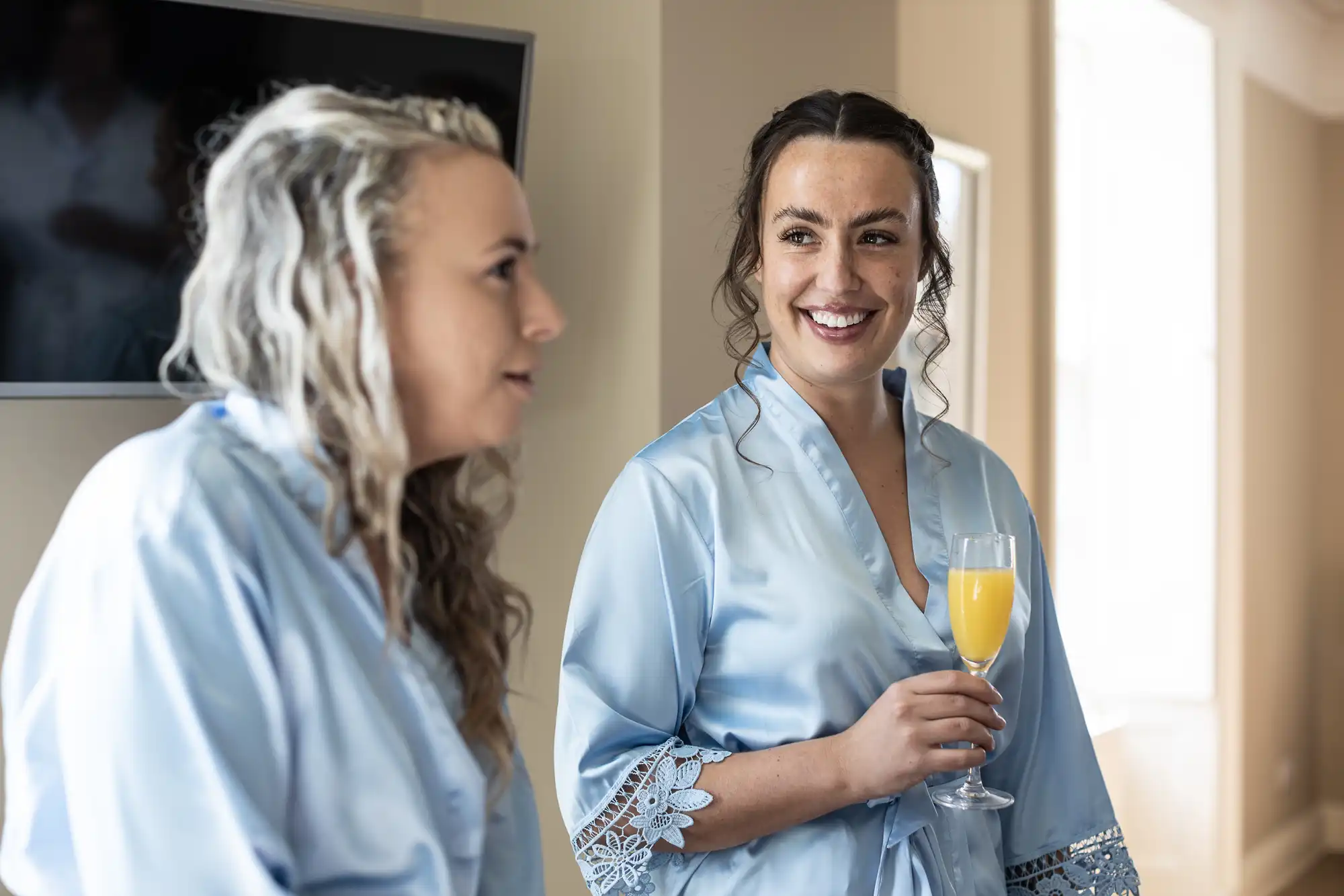 Two women in light blue robes, one holding a drink, engage in conversation in a well-lit room with a TV in the background.