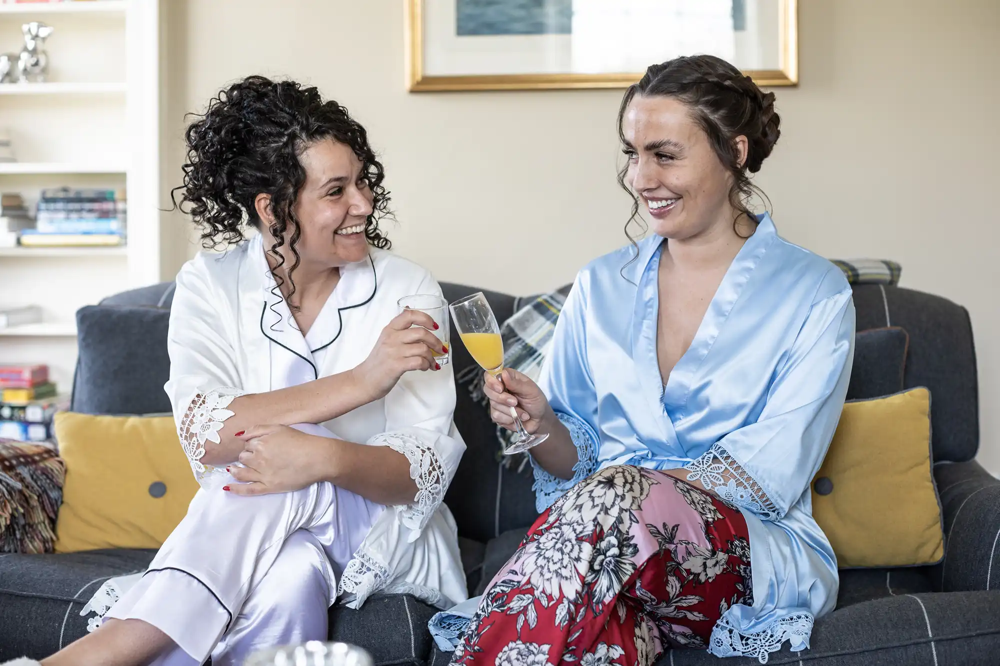 Two women in pajamas and robes sit on a couch, smiling and toasting with champagne flutes filled with an orange beverage.