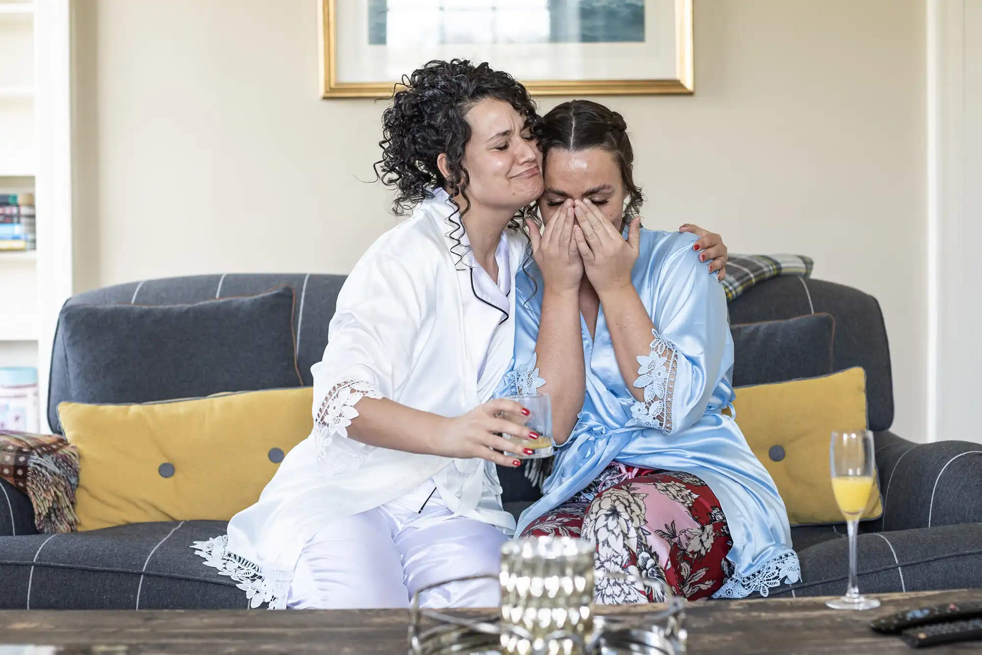 Two people in matching loungewear sit on a couch. One person comforts the other by holding them close while the other covers their face with their hands. Drinks are on the table in front of them.