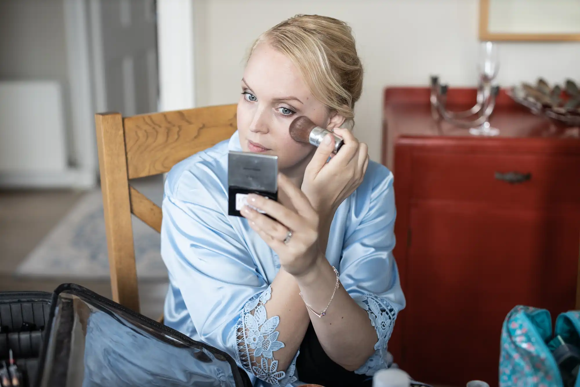 A person wearing a blue robe applies makeup with a brush while looking into a compact mirror.