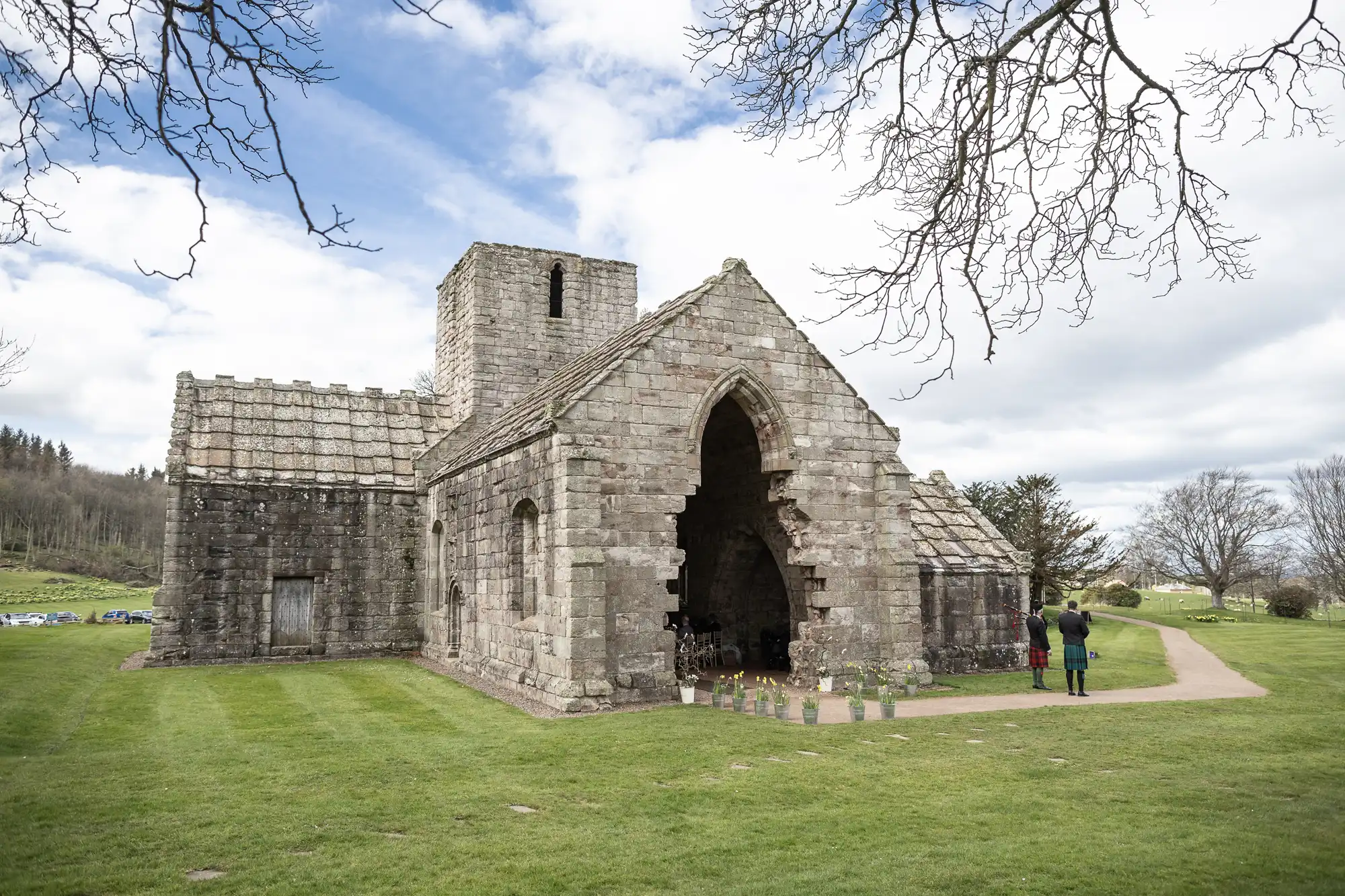 A historic stone church with a central tower and arched entrance, set on a grassy area with a few people standing nearby under a cloudy sky.