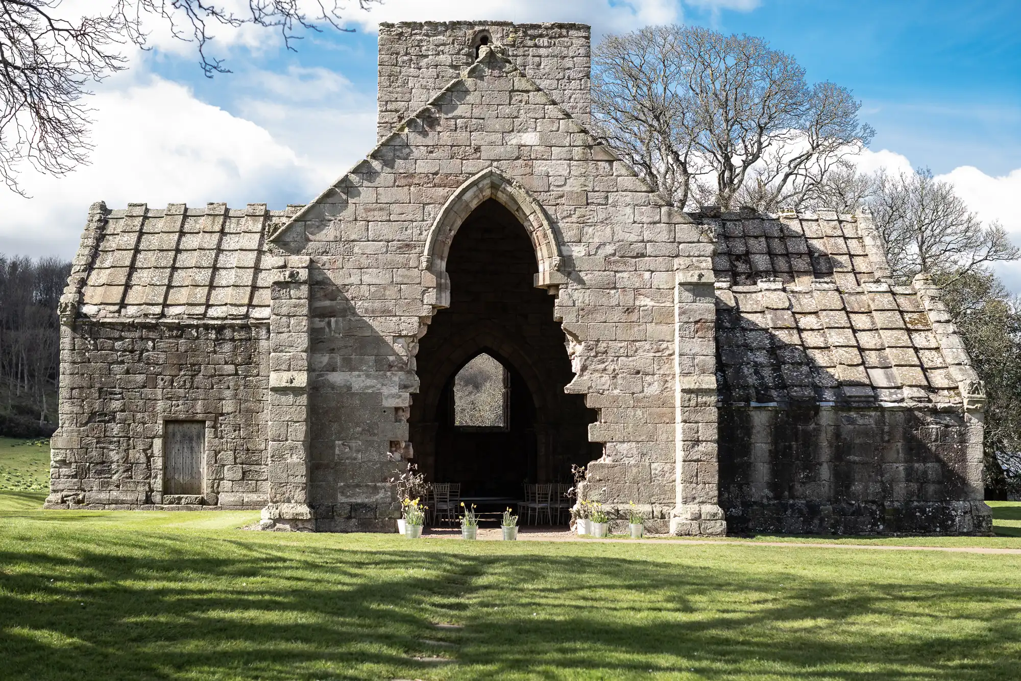 A stone building with a Gothic-style arch stands on a well-manicured grassy lawn under a blue sky with scattered clouds. The structure appears to be a historic or abandoned church or chapel.