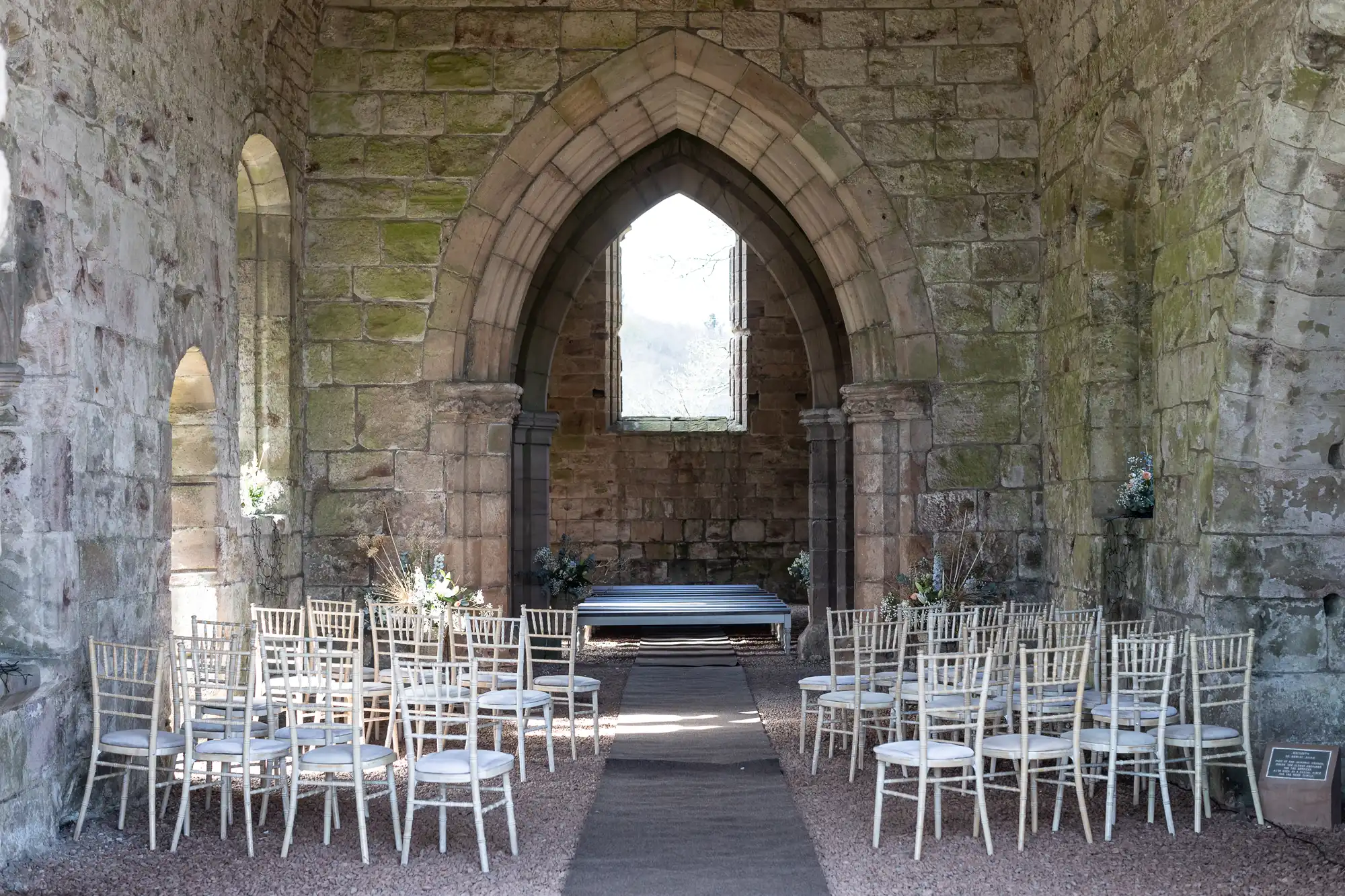 An empty stone-walled chapel with rows of white chairs arranged for a ceremony, facing a small stage under an arched window.