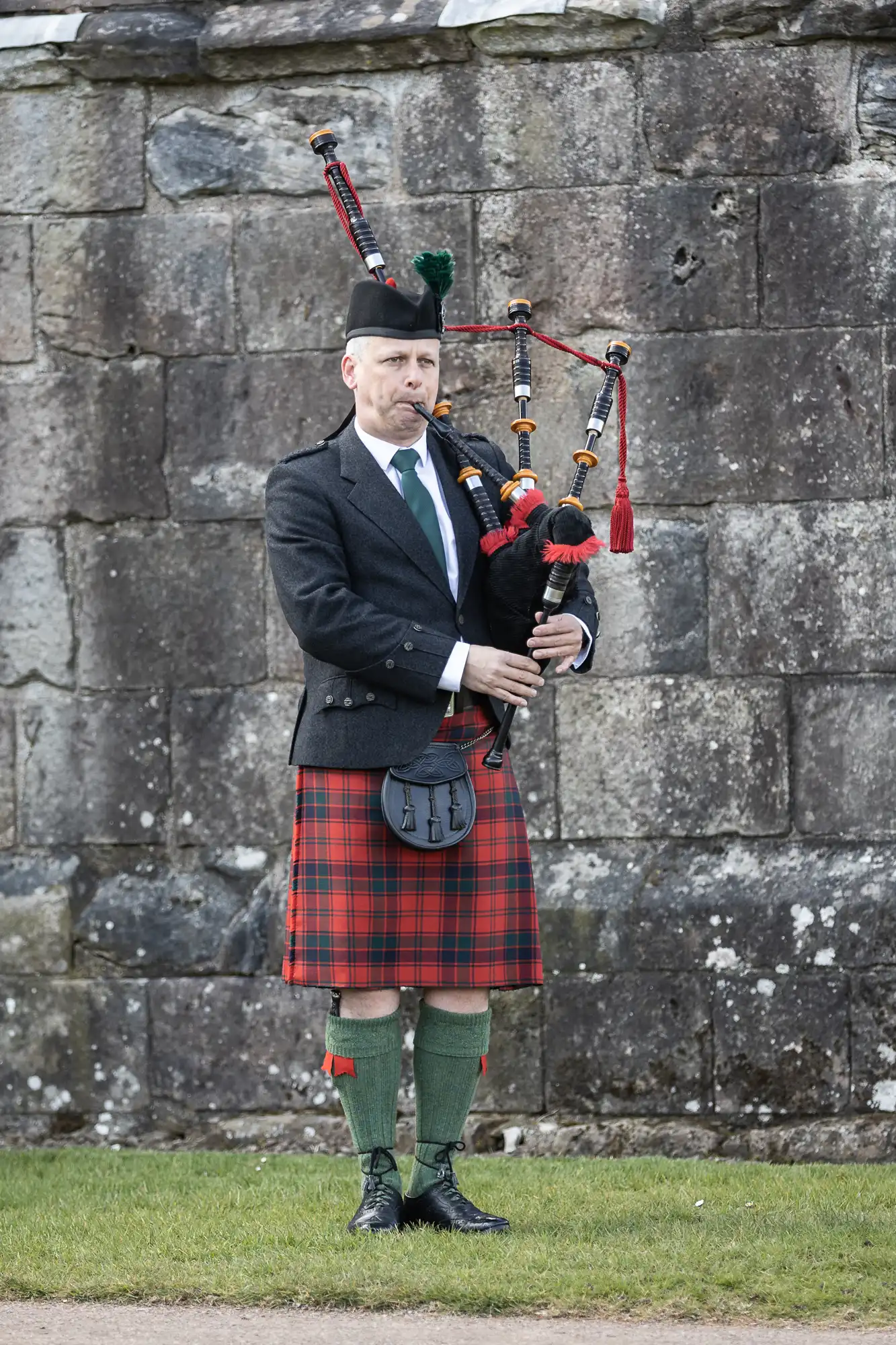 A man wearing a traditional Scottish kilt and a sporran is playing the bagpipes in front of a stone wall.