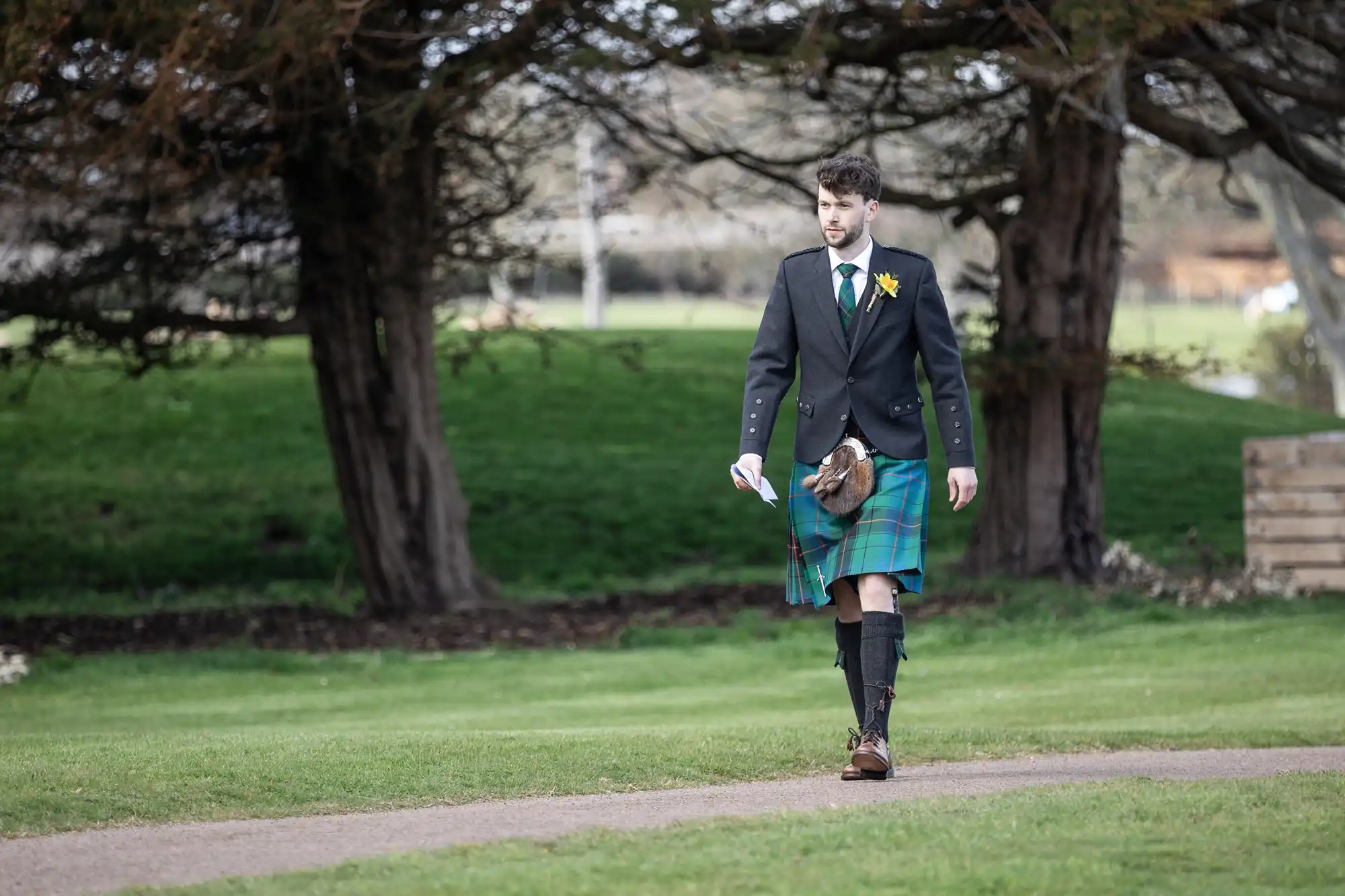 A man wearing a green kilt, black jacket, and knee-high socks walks on a path in a grassy area with trees in the background. He holds a small object in one hand.