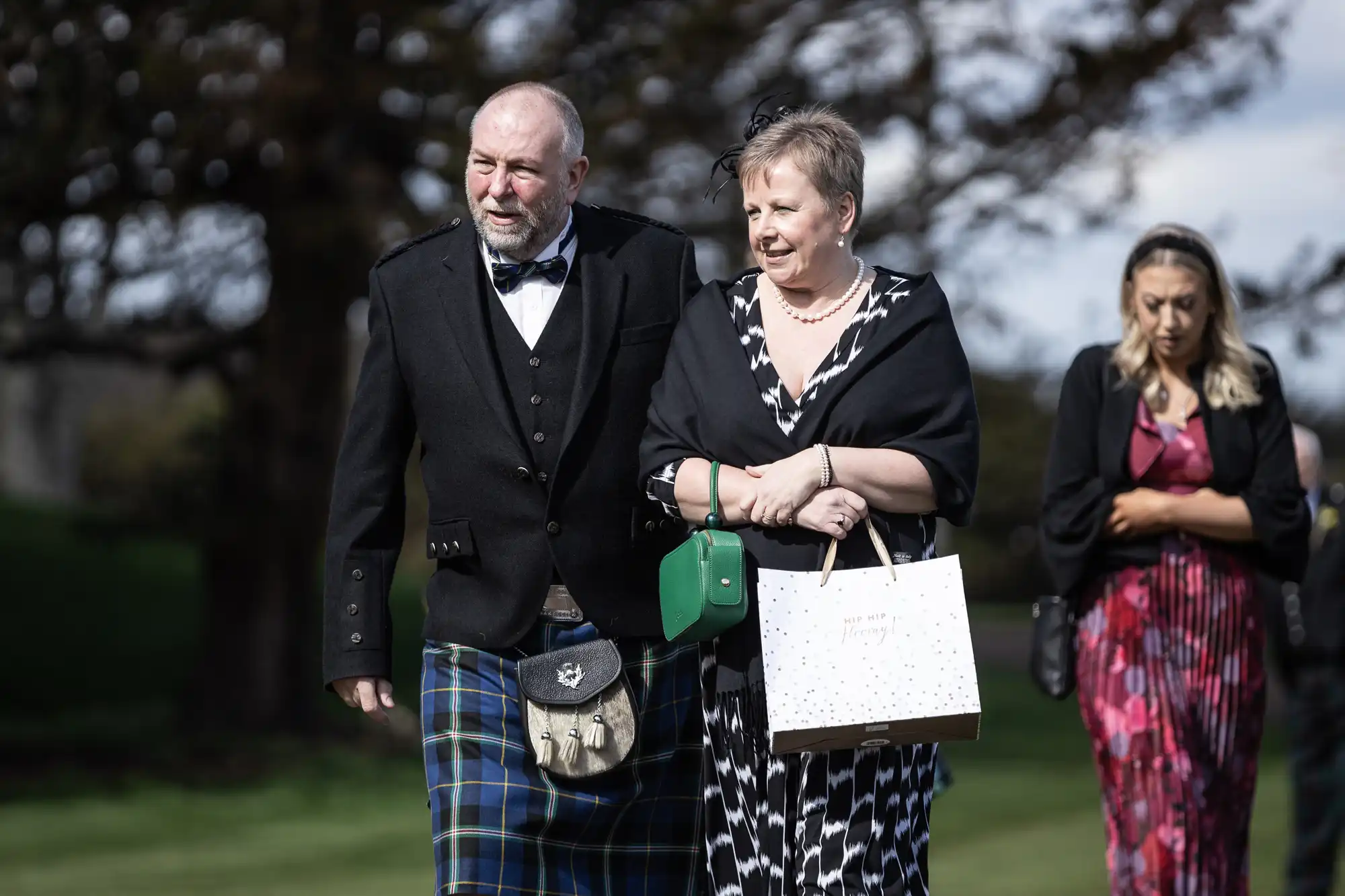 A man in a kilt and a woman holding a gift bag walk together outdoors, followed by another woman in a red dress with a black jacket.