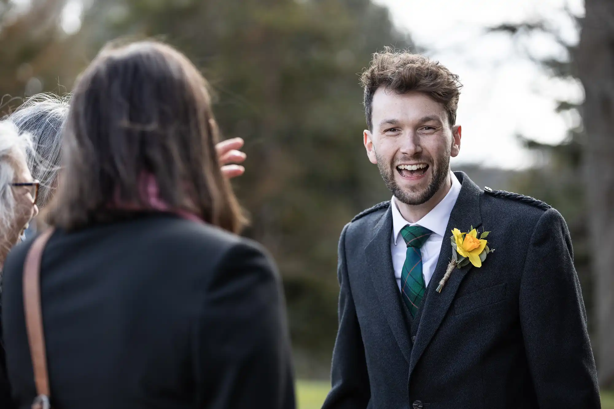 A man in a suit with a yellow flower boutonnière is smiling and talking to two people outdoors.