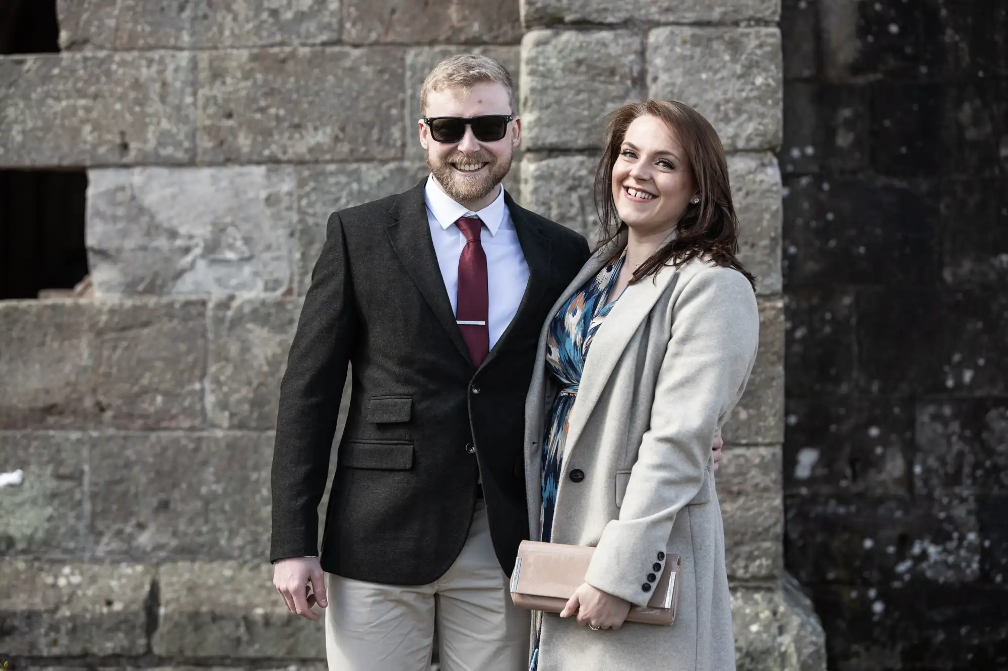 A man in a dark blazer and a woman in a light coat stand smiling in front of an aged stone wall. The man wears sunglasses and holds a beverage, while the woman carries a clutch.