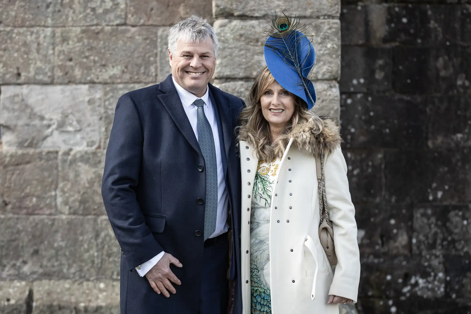 A man and woman stand together smiling. The man is wearing a dark suit and the woman is in a white coat with a blue hat adorned with a peacock feather. They are outdoors in front of a stone wall.