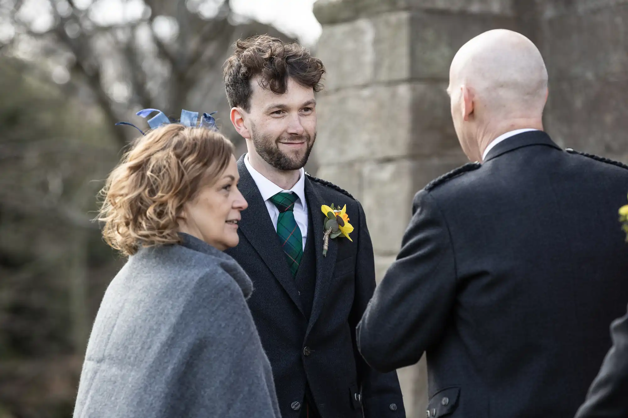 Three people are having a conversation outside near an old stone structure. The man in the middle is wearing a suit with flowers pinned on his lapel.