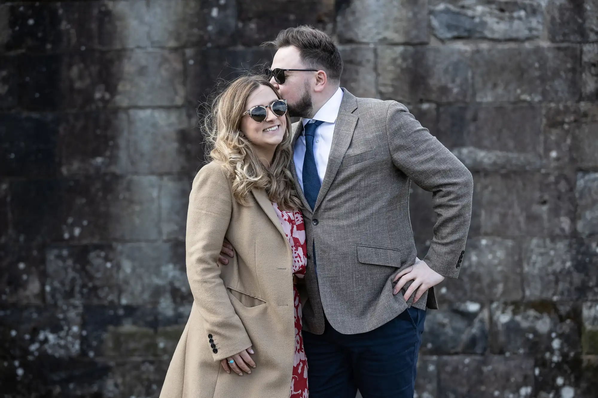 A man in a brown blazer and sunglasses kisses a smiling woman in a beige coat and sunglasses on the head, standing in front of a stone wall.