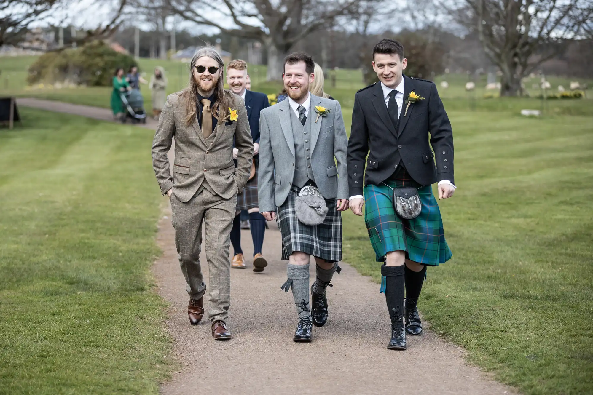 Four men in suits and kilts walk down a path in a park, smiling and talking. The men are wearing traditional Scottish attire, including kilts and sporrans, and some have daffodil flowers pinned to their jackets.