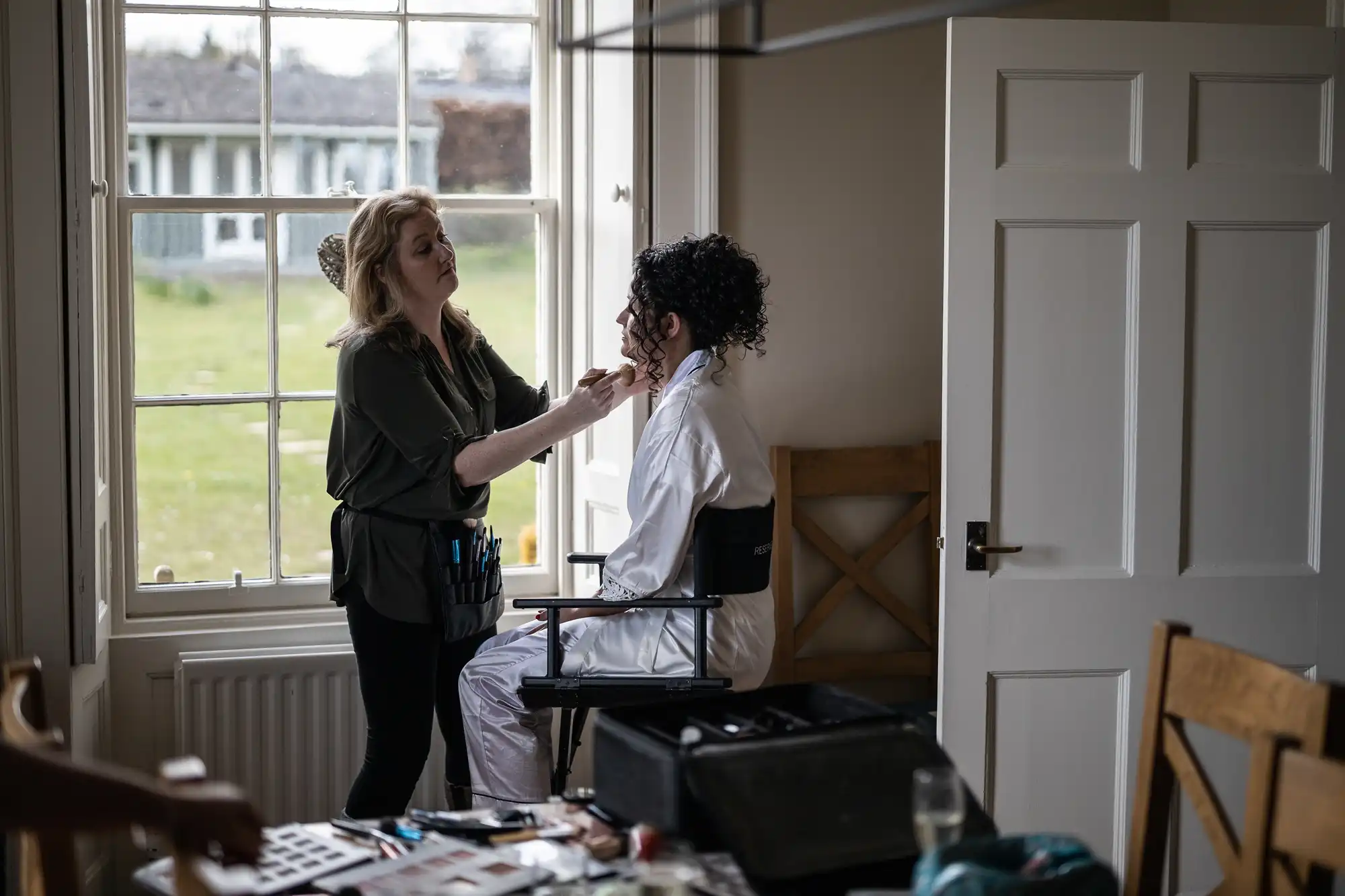 A makeup artist applies makeup to a seated individual by a window in a well-lit room with a door and various beauty tools on a table.