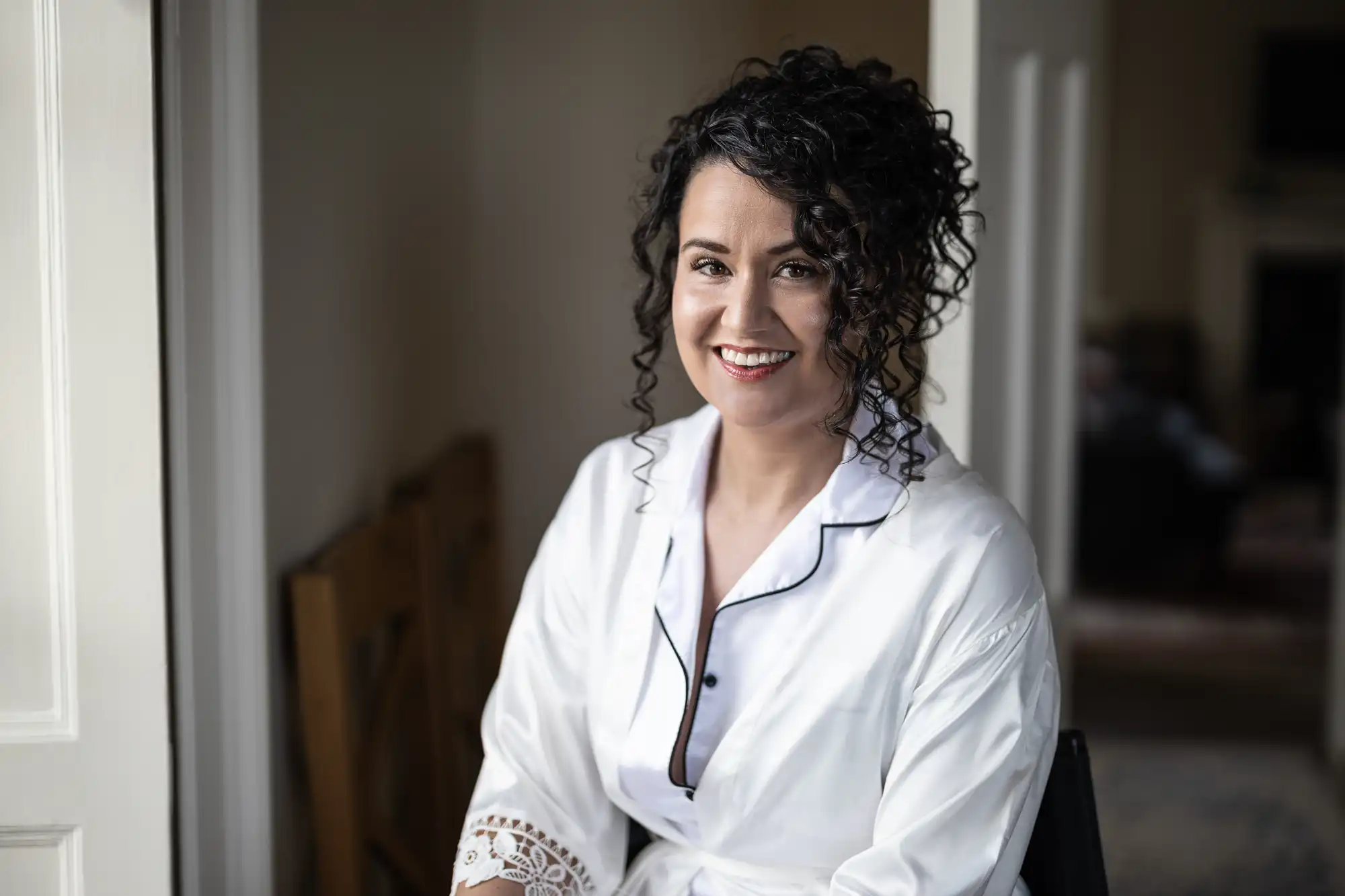 A woman with curly hair, wearing a white robe, smiles at the camera while sitting indoors.