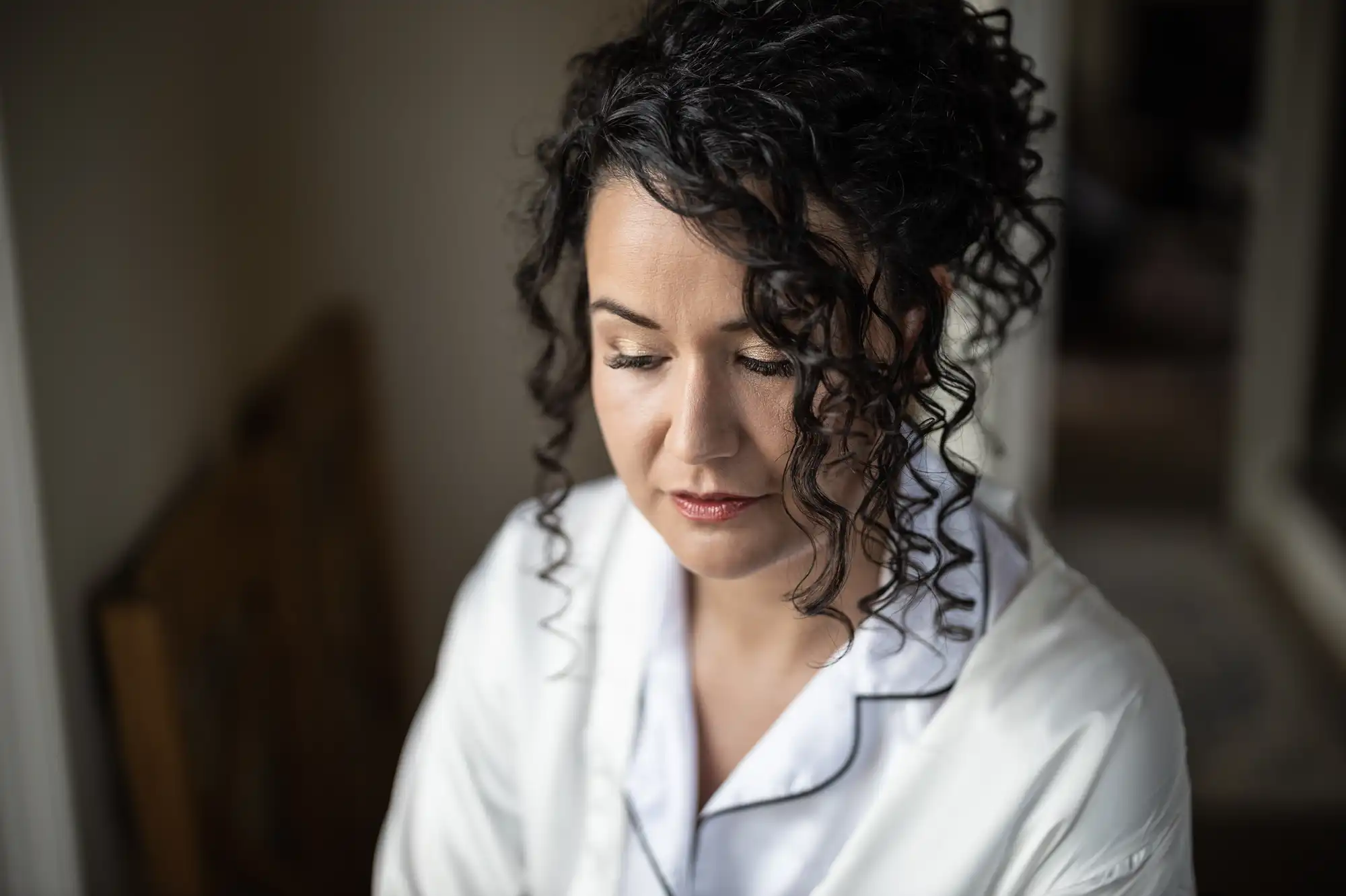 A woman with curly dark hair, wearing a white garment with black piping, looks down with her eyes closed.