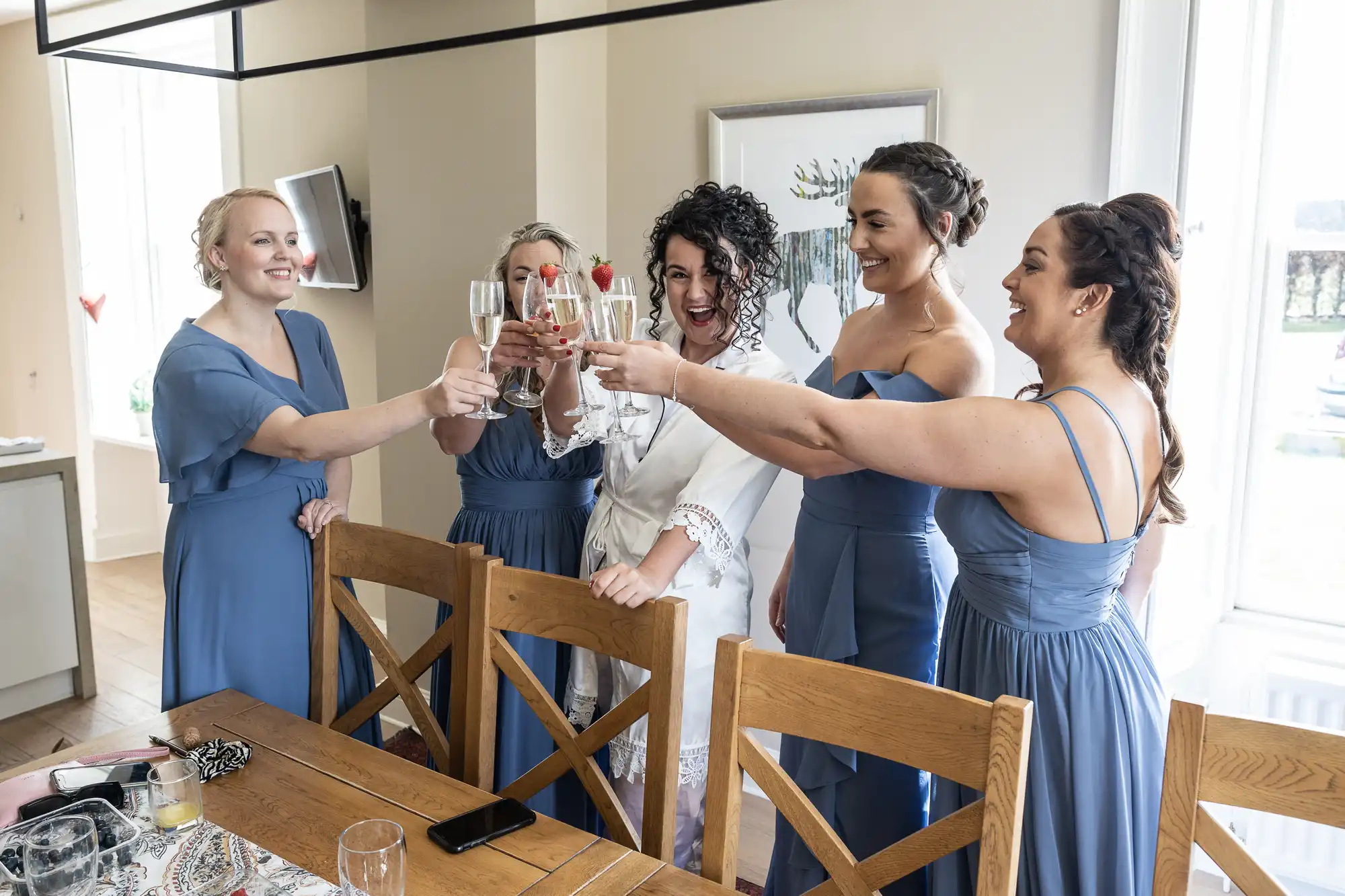 A bride and four bridesmaids in blue dresses smile and raise champagne glasses for a toast in a bright room with wooden furniture.