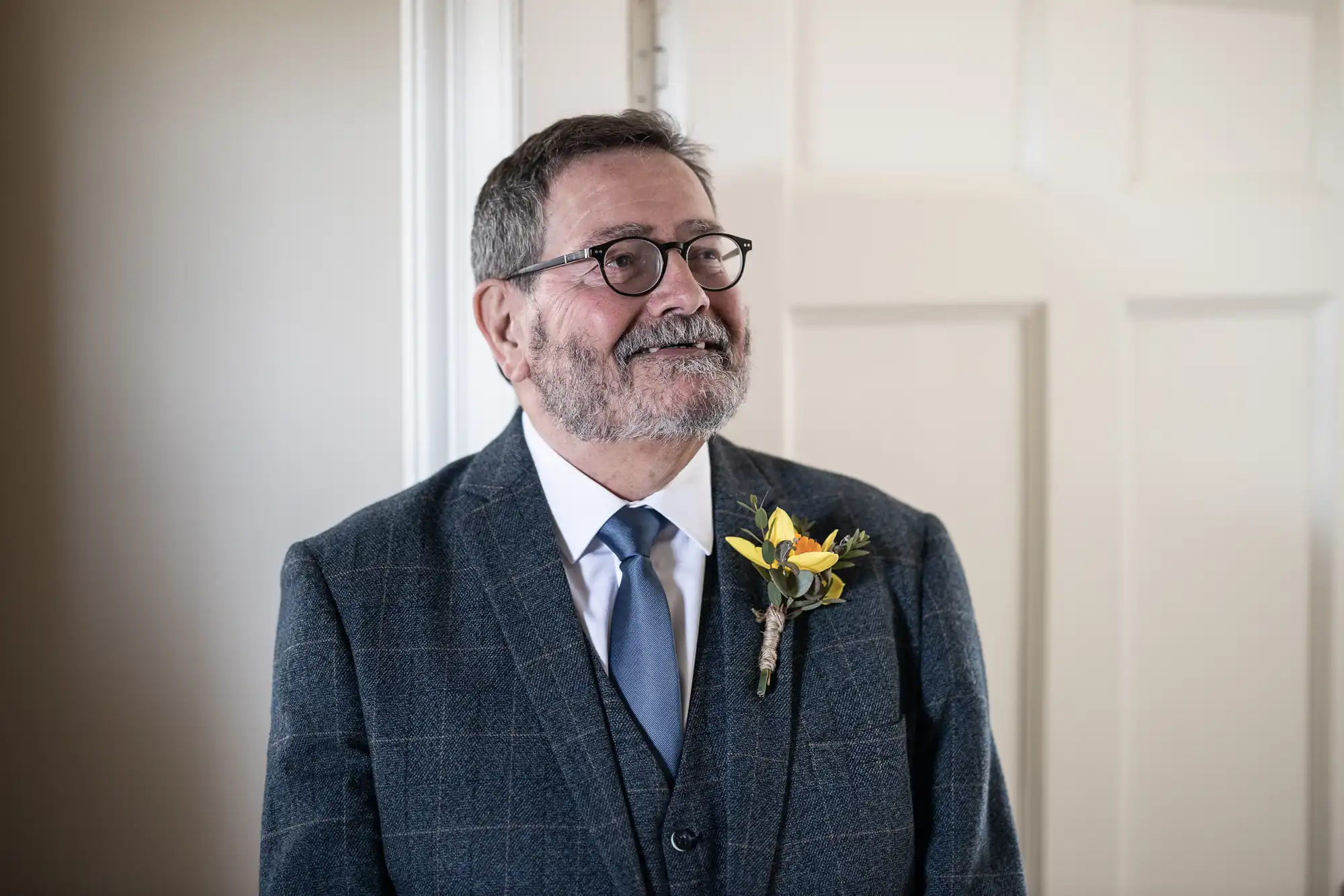 A man with glasses and a beard, wearing a checkered suit with a blue tie and a yellow flower boutonniere, stands in front of a white door.