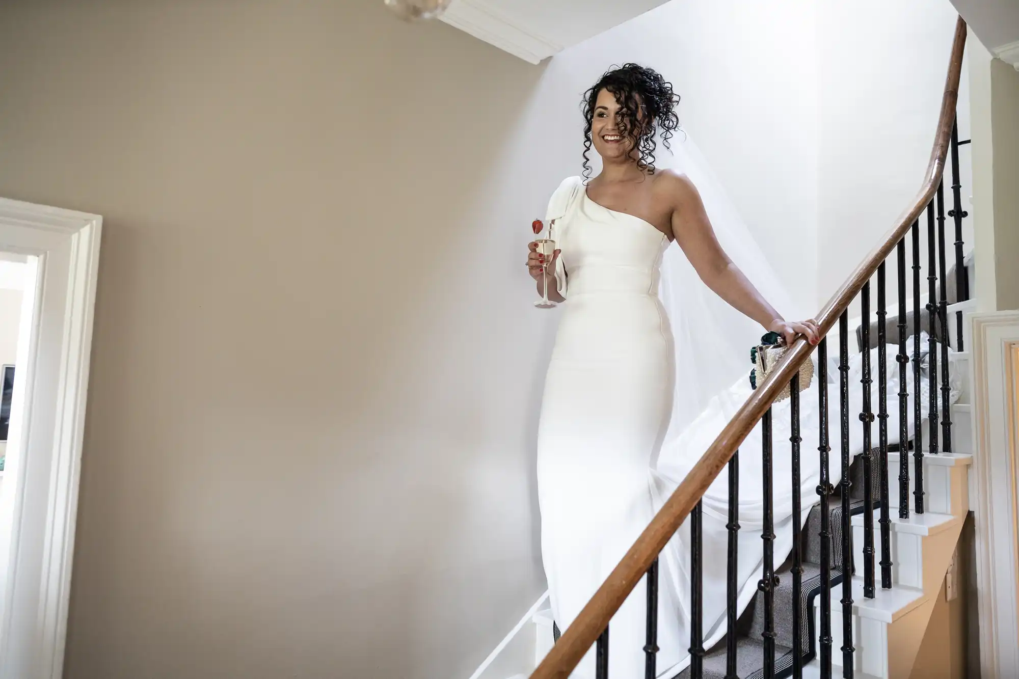 A bride in a white dress descends a staircase holding a glass of champagne.