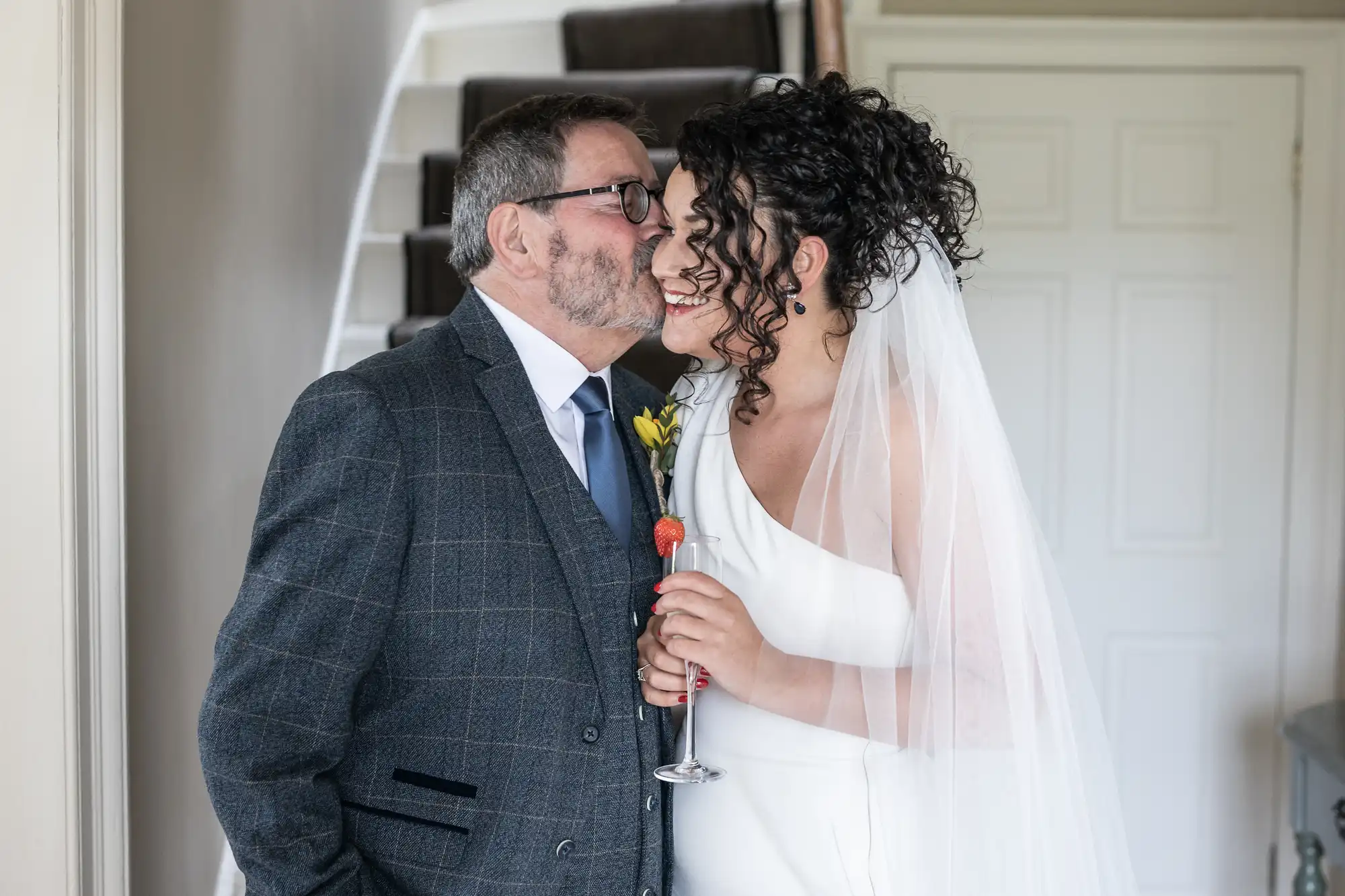 A couple, dressed in wedding attire, is standing closely. The man, wearing a suit, is kissing the bride on the cheek. The bride is in a white gown with a veil, holding a bouquet.