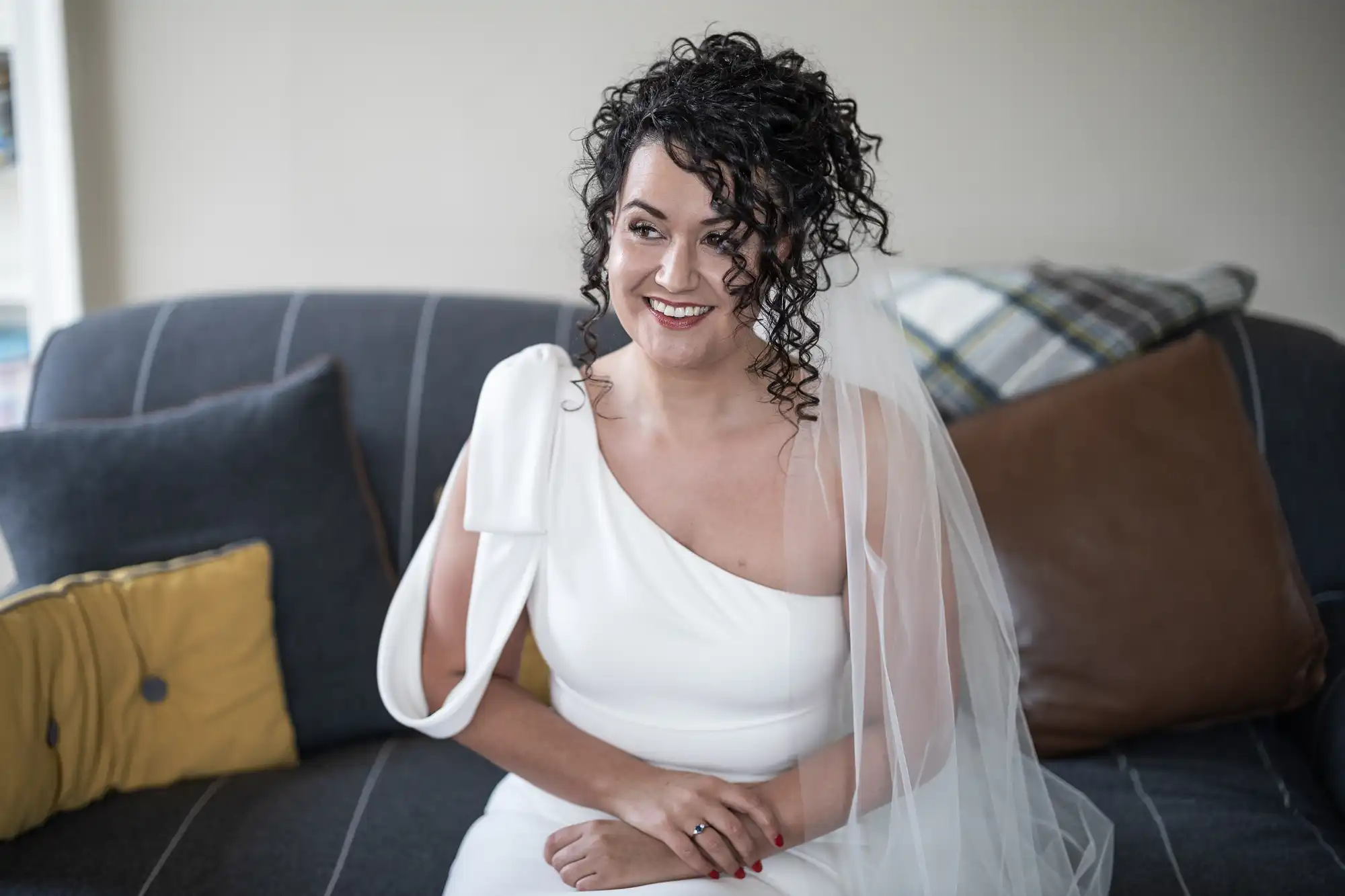 A woman with curly hair smiles while seated on a gray couch, dressed in a white gown and veil.