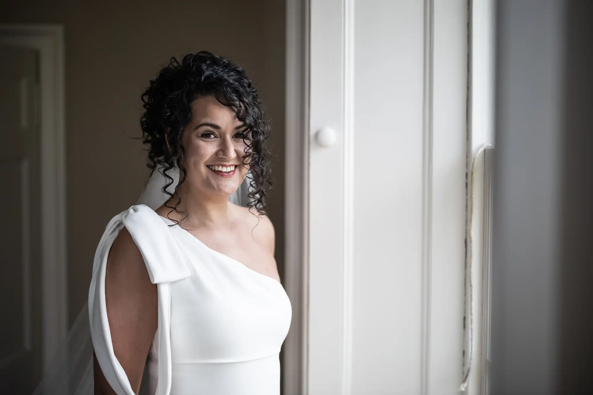 A woman with curly hair, wearing a white off-shoulder dress, smiles while standing near a window indoors.