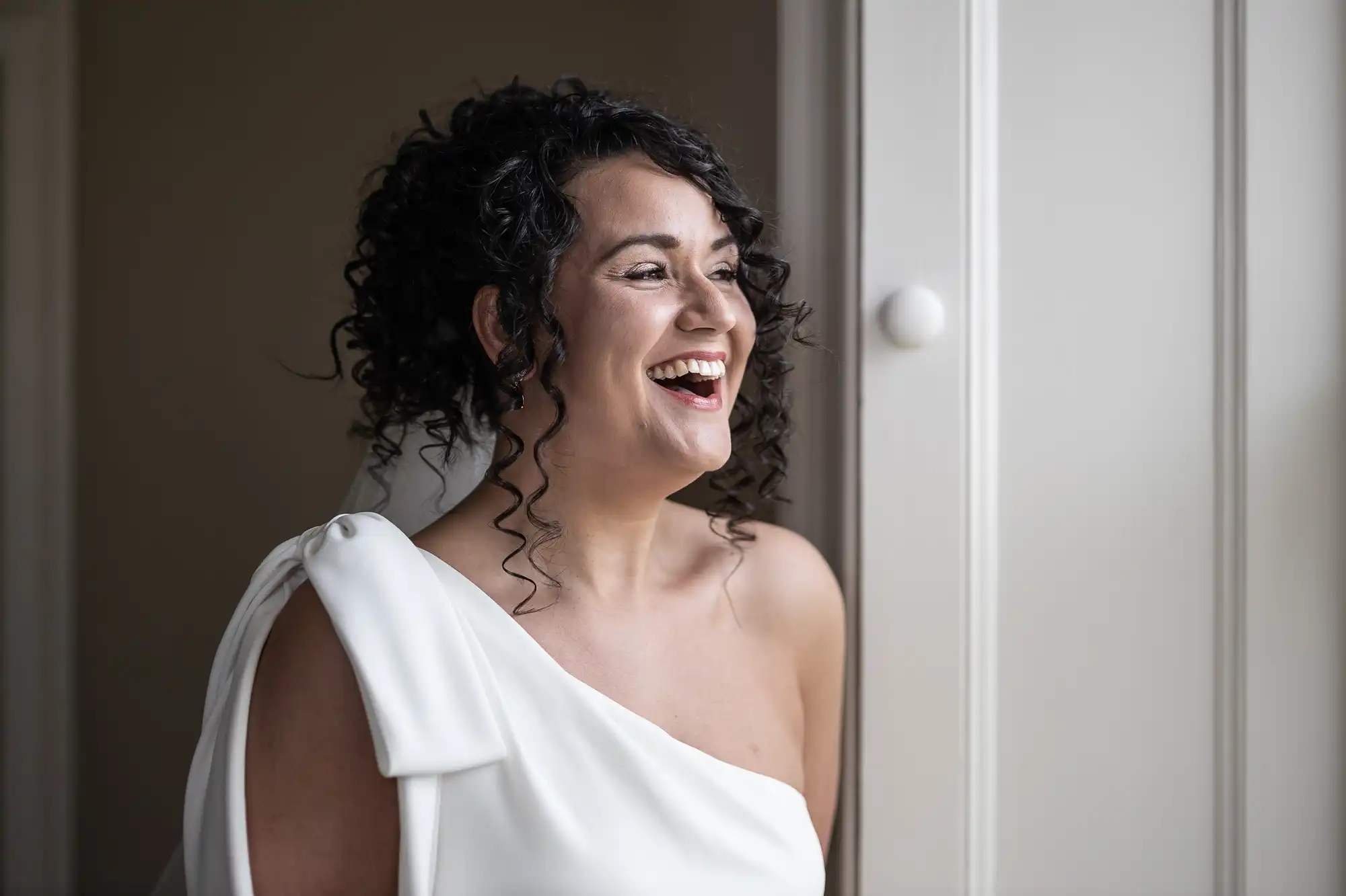 A woman with curly hair and a white one-shoulder dress smiles while looking to the side near a window.