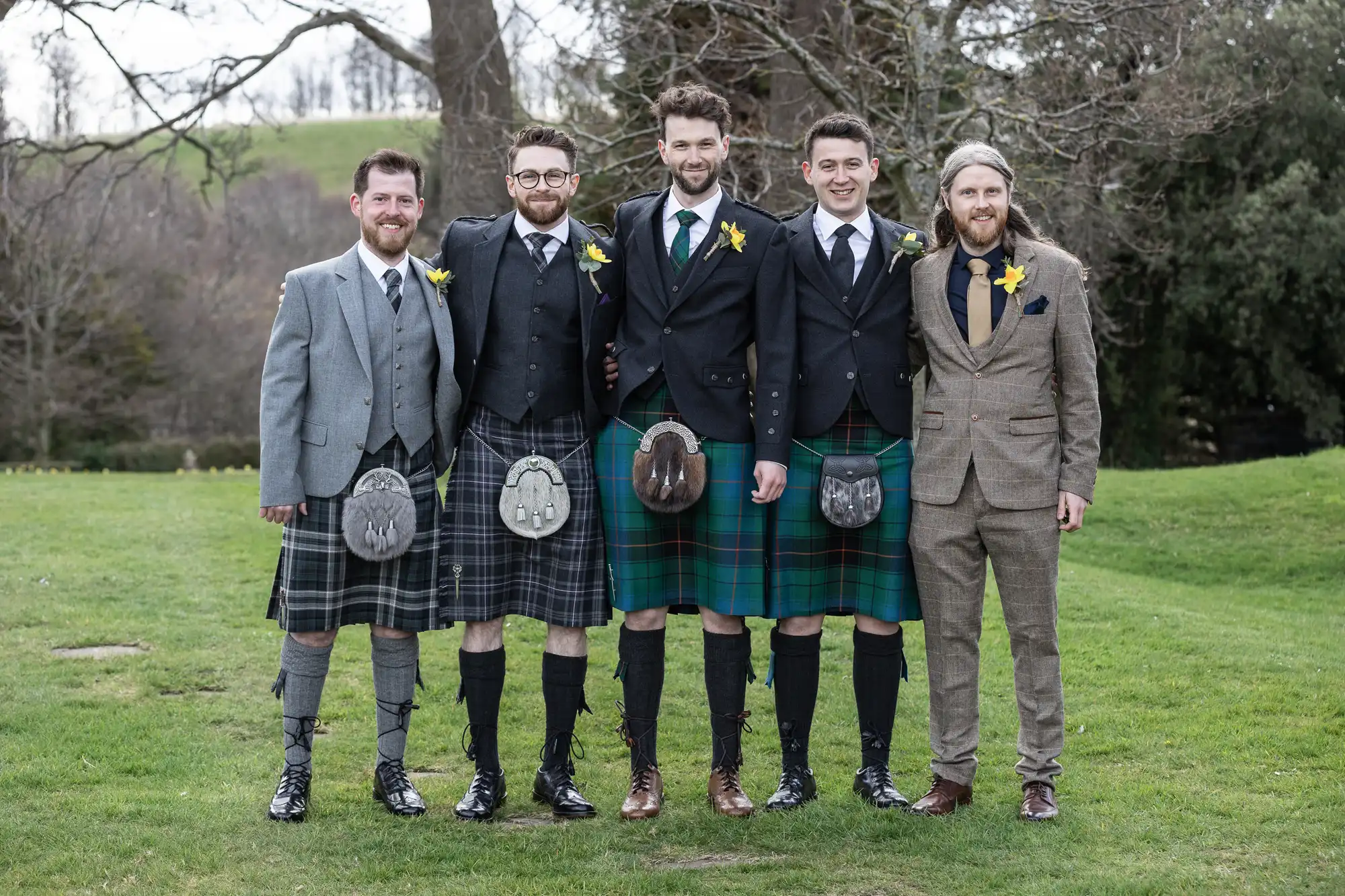 Five men in formal attire, three wearing kilts and two in suits, standing on a grassy outdoor area with trees in the background.