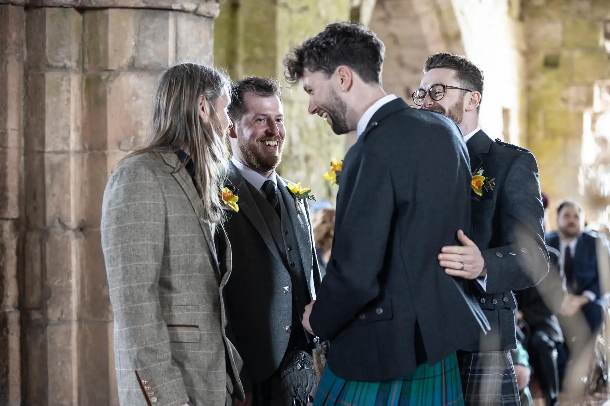 Four men, one in a kilt and the others in suits, stand together laughing in a stone building.