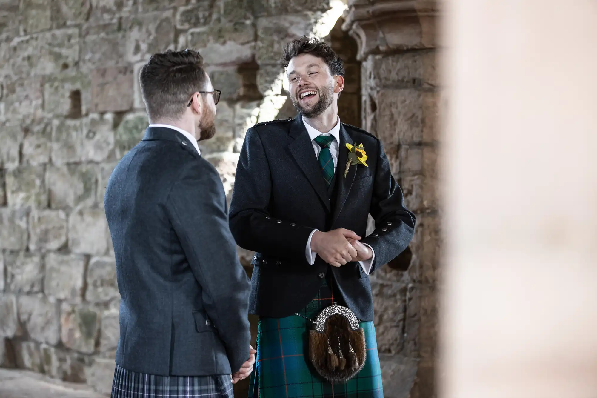 Two men wearing kilts and blazers share a laugh inside a stone building. One man has a yellow flower on his lapel and they both have beards and glasses.