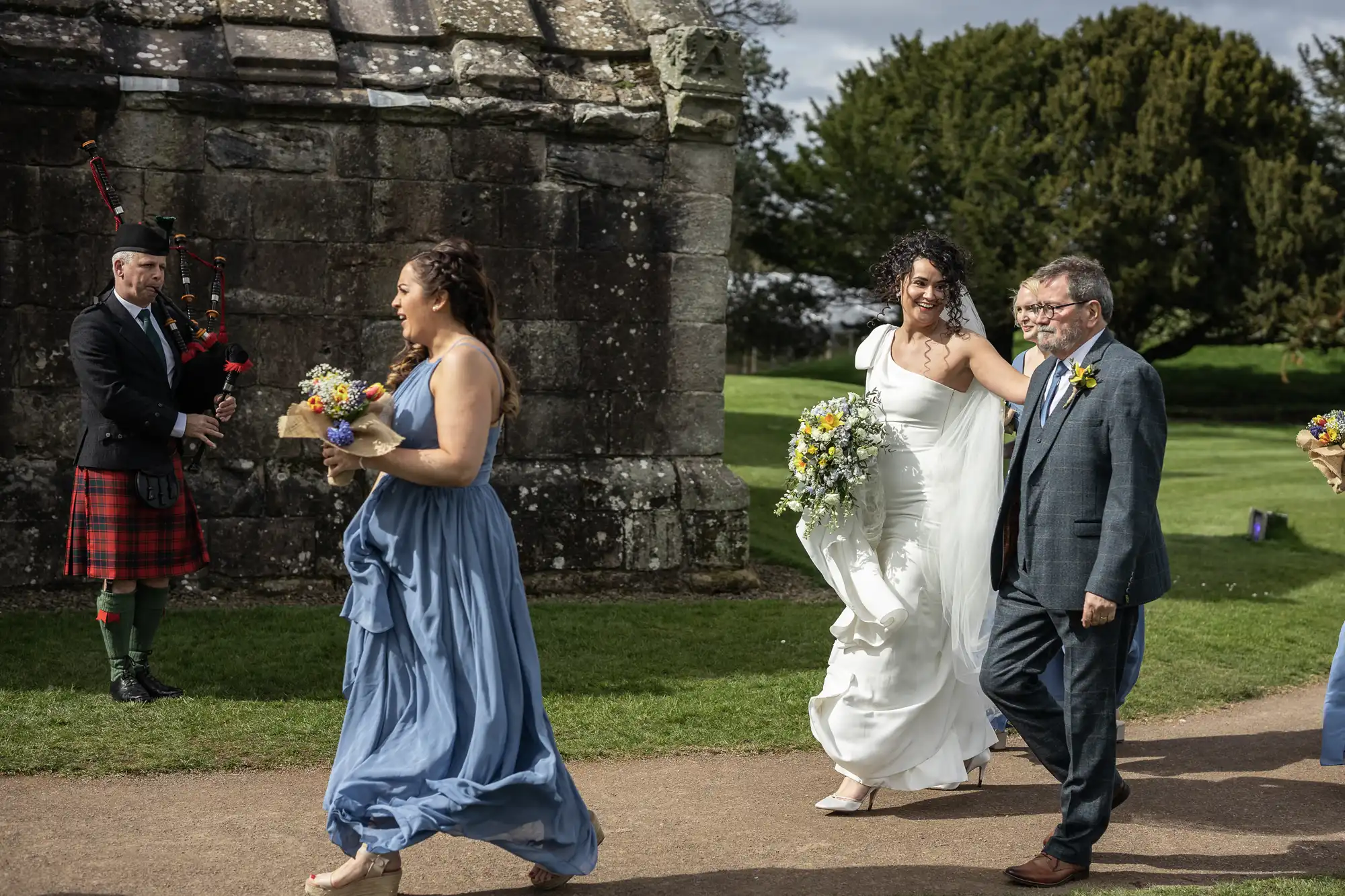 Bride holding a bouquet and arm in arm with a man walks behind a bridesmaid in a blue dress, with a bagpiper in a kilt playing nearby, outdoors near an old stone structure.