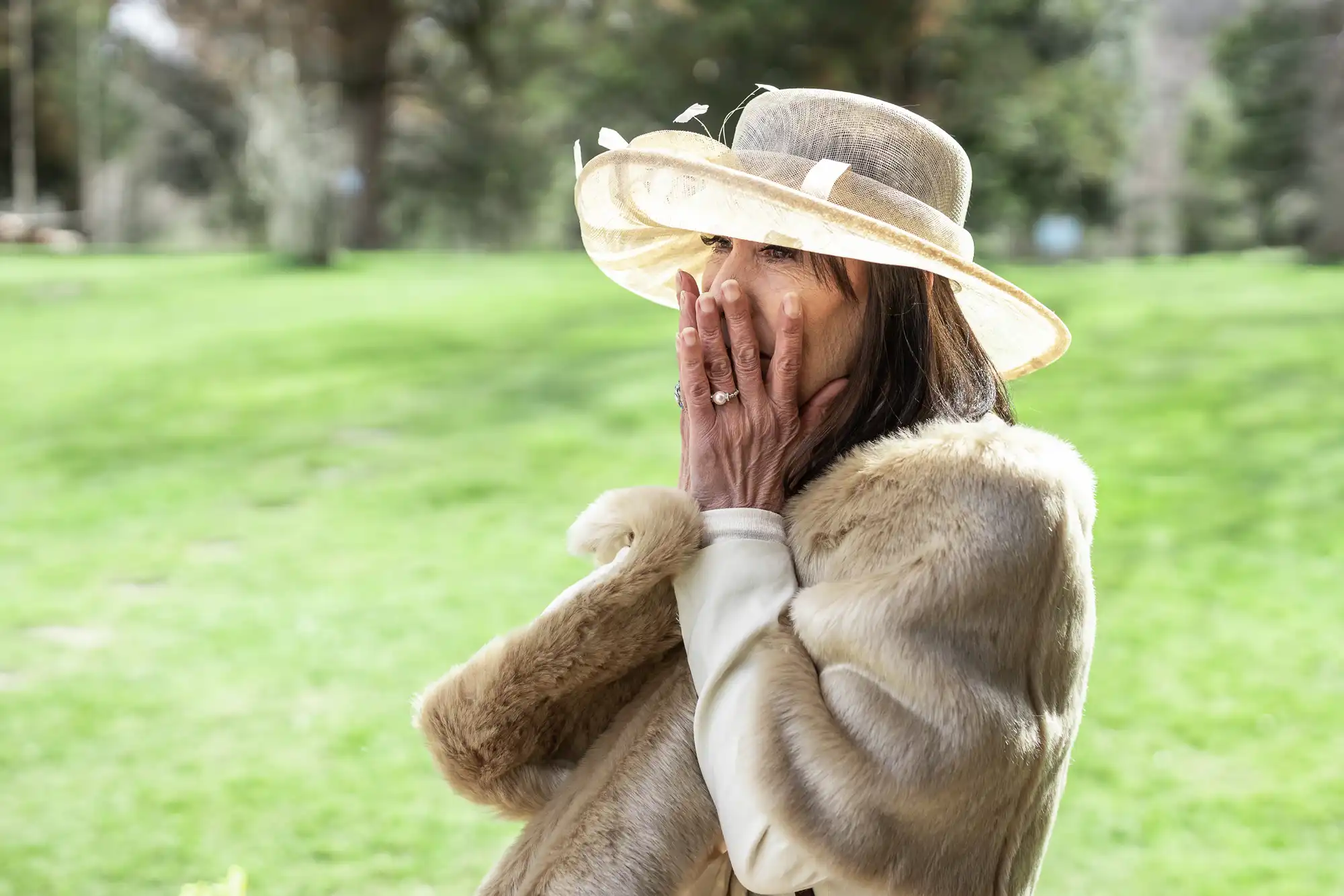 A woman wearing a wide-brimmed hat and fur wrap covers her mouth with both hands while standing outdoors on a sunny day.