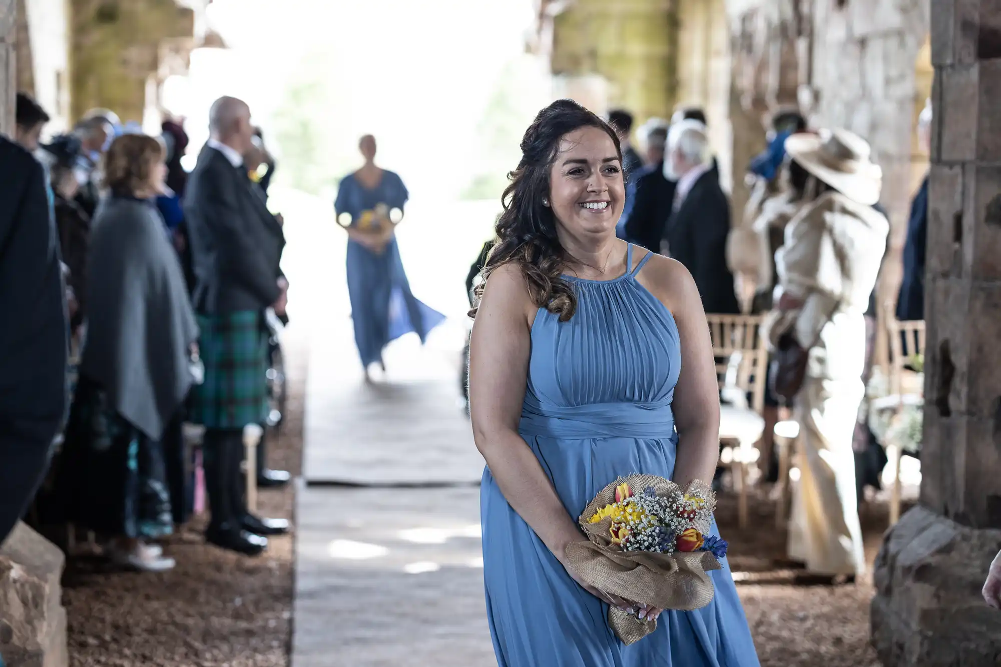 A woman in a blue dress walks down an aisle holding a bouquet of flowers at an outdoor event. People are seated on both sides, watching her.