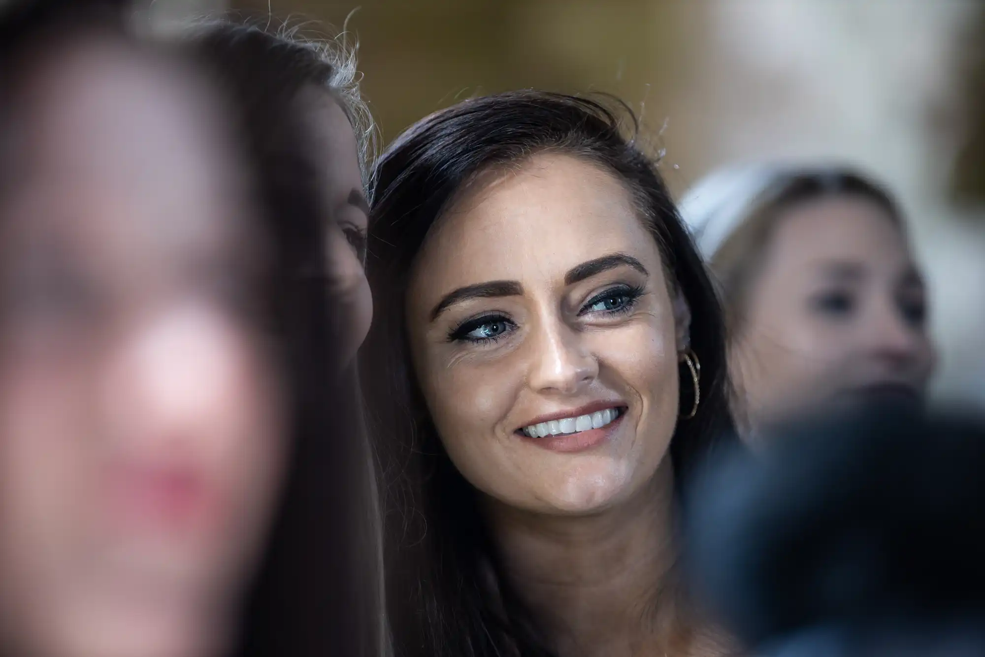 A woman with long dark hair, wearing hoop earrings and smiling, is surrounded by other people in a blurred background.