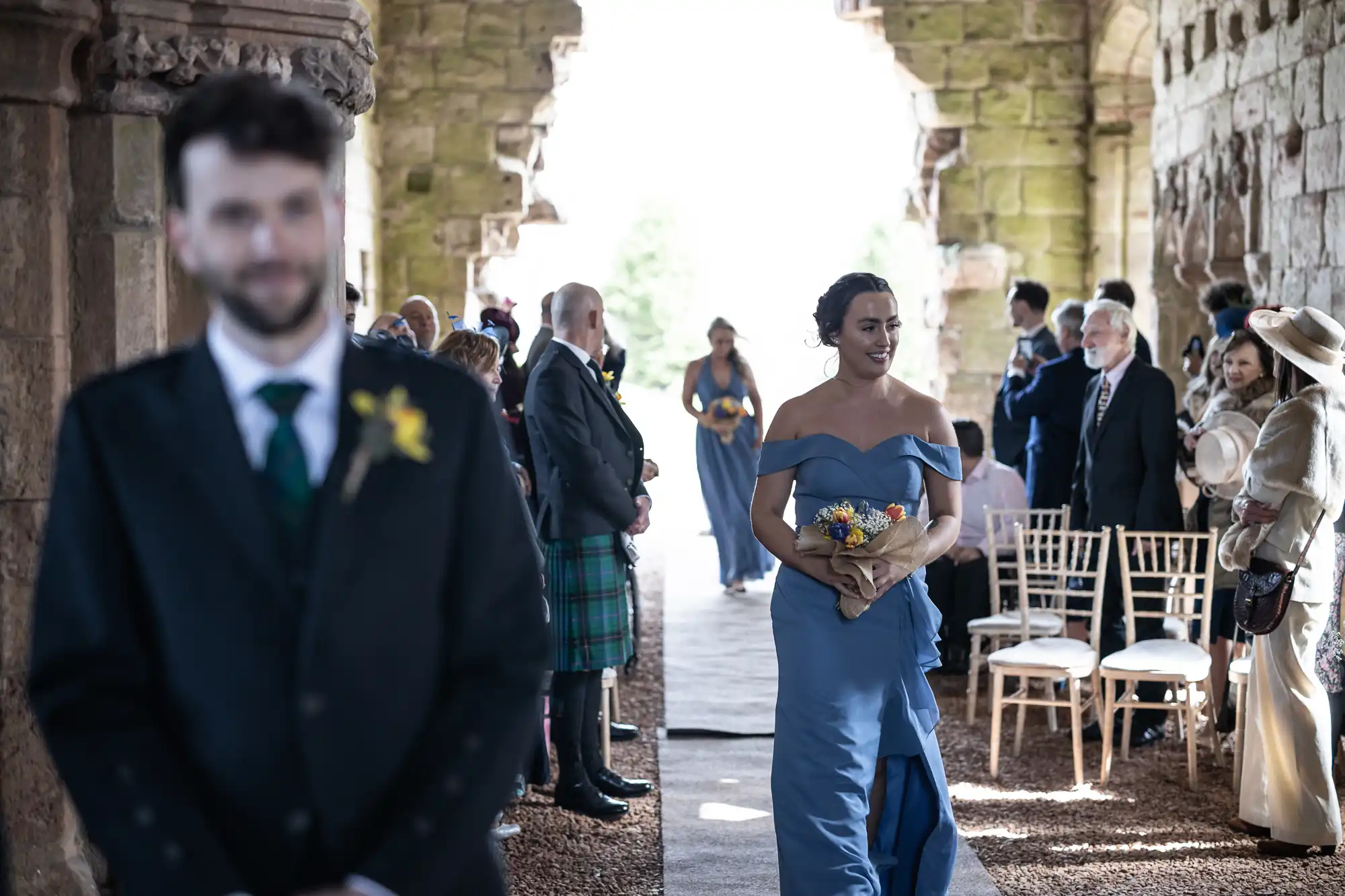 A bridesmaid in a blue dress walks down the aisle holding a bouquet, with guests seated on either side and a man in a suit standing in the foreground at an indoor ceremony venue.