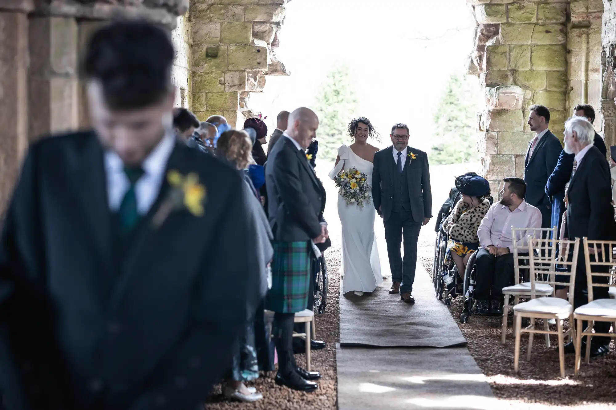 A bride walks down the aisle with a man, likely her father, during a wedding ceremony. Guests are seated on both sides, some standing, and one person is in a wheelchair.