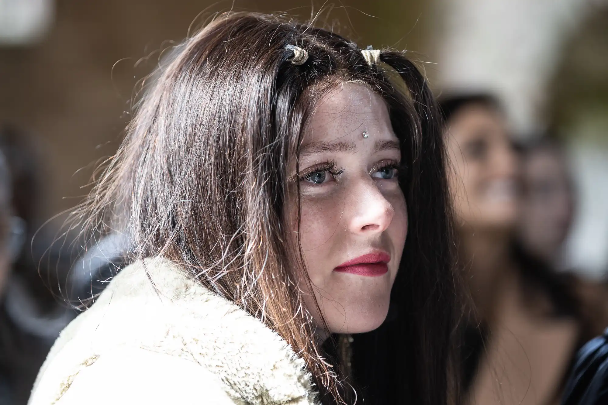 Close-up of a person with long brown hair and decorative hair accessories, wearing a light fur coat. The background is blurred with several people slightly visible.