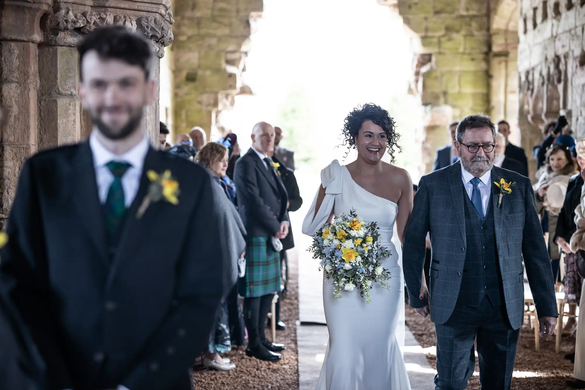 Bride in white dress holding yellow flower bouquet walks down the aisle with older man in a stone building, people seated and standing on either side, blurred man in foreground.