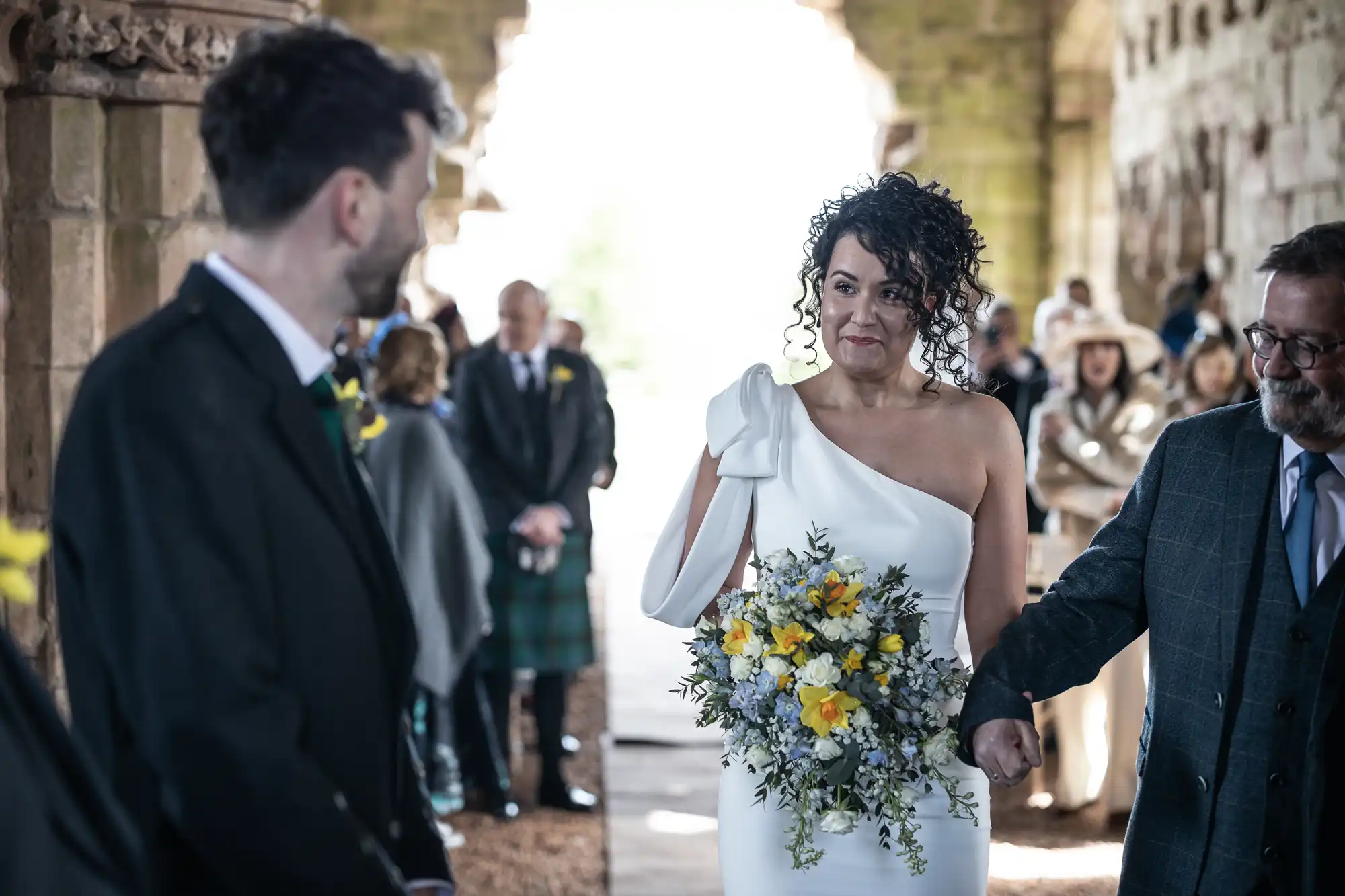 A bride holding a bouquet of yellow and white flowers walks down the aisle with an older man in a formal setting, approaching a groom in a black suit. Guests are seated in the background.