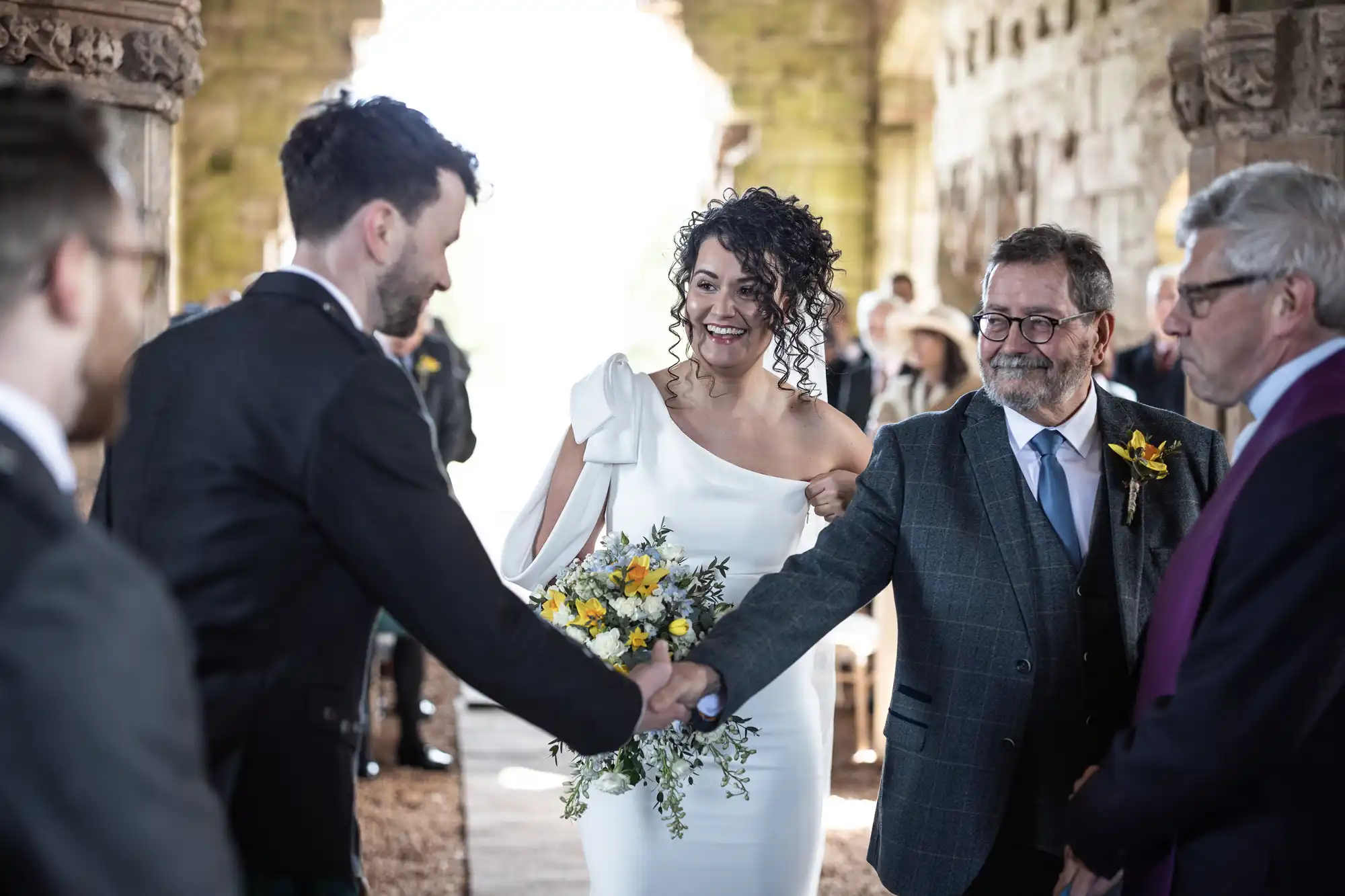 Bride in a white dress holding a bouquet, standing with an older man in a suit, smiles as another man shakes hands with a groom in a dark suit in an ornate, stone-walled venue.