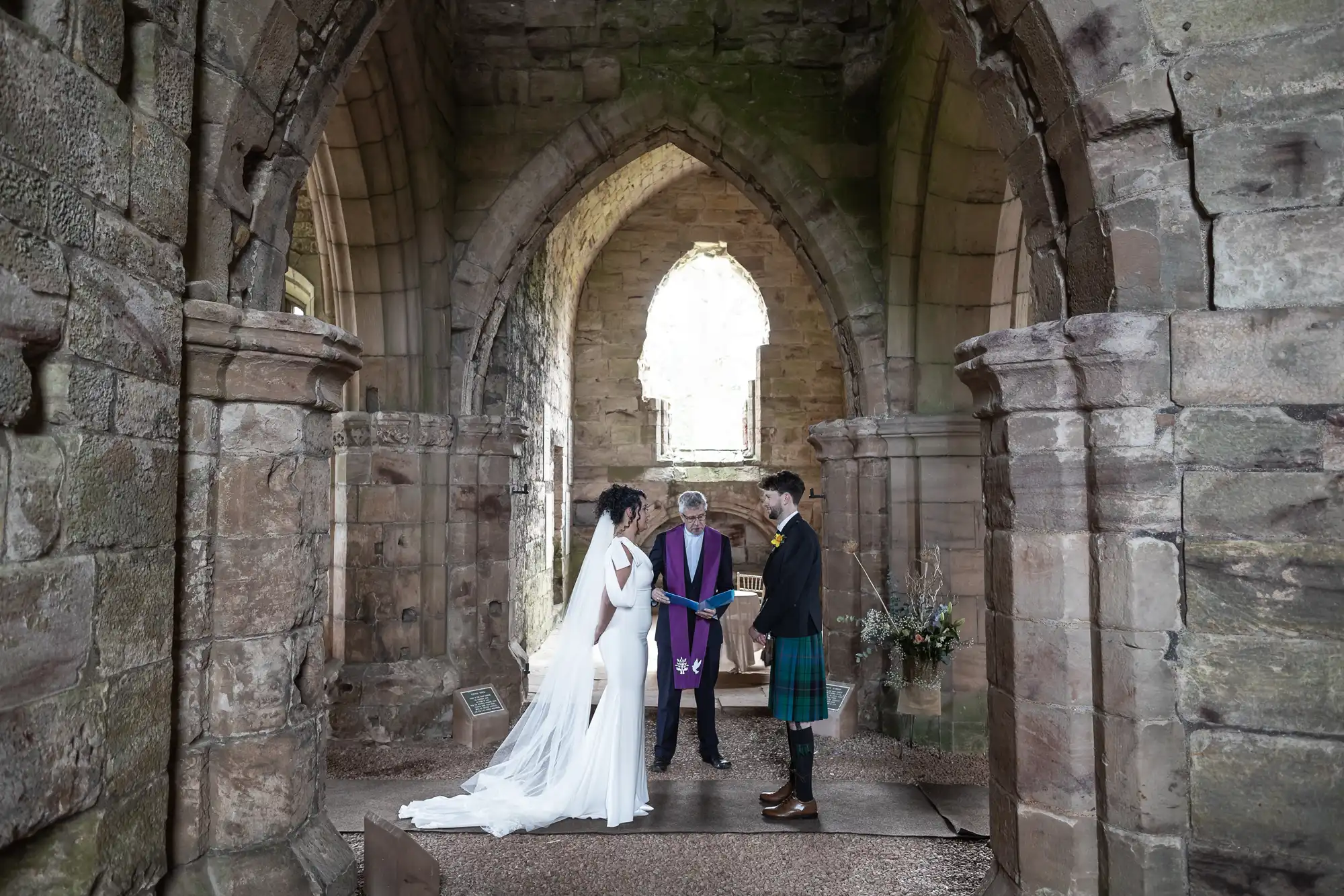 A wedding ceremony taking place inside an ancient stone building. The couple stands in front of an officiant, with the arches and stonework visible in the background.