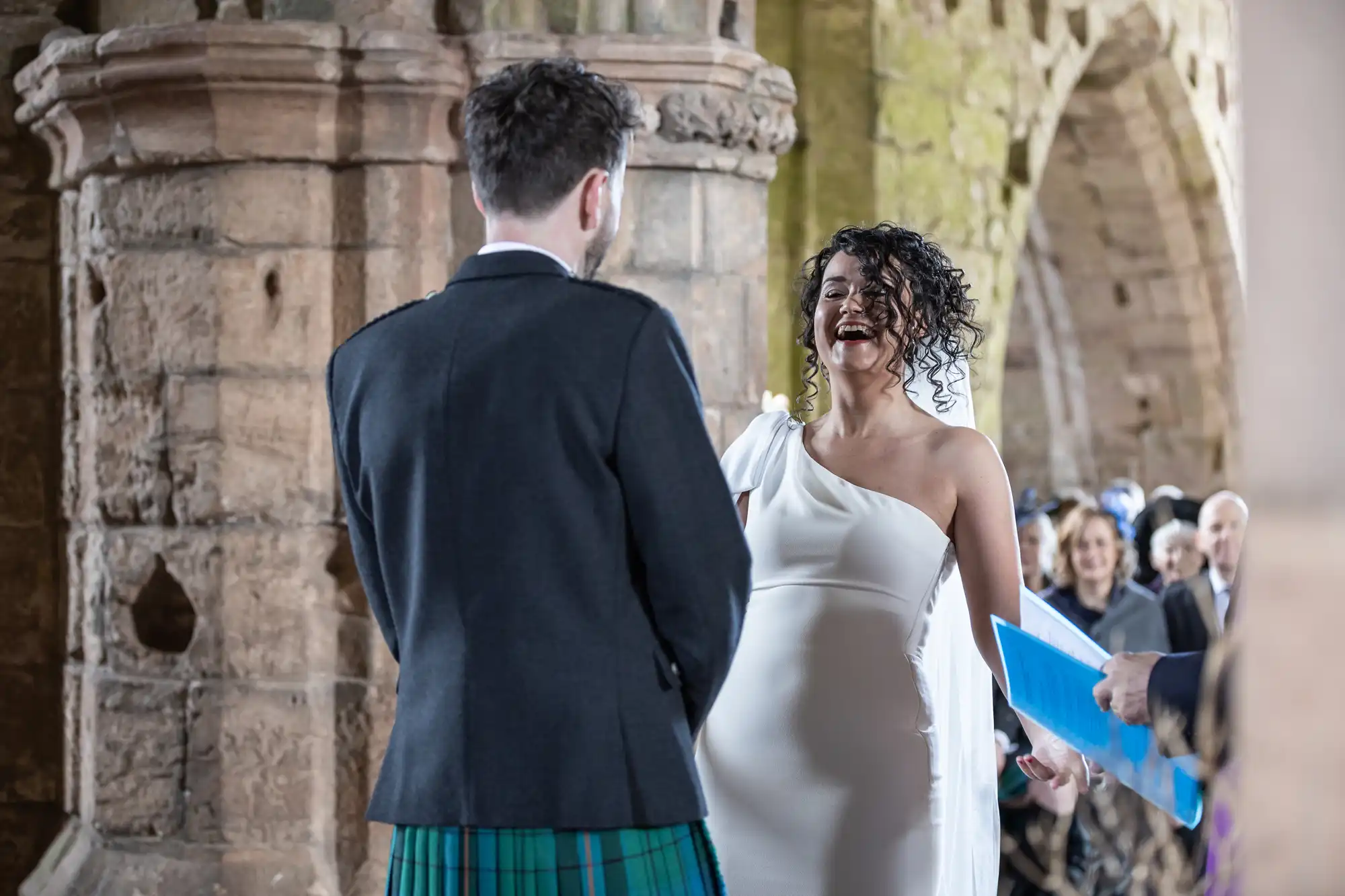 A couple stands together, smiling during their wedding ceremony held in a historic stone building, with guests seated in the background. The bride wears a white dress, and the groom wears traditional attire.