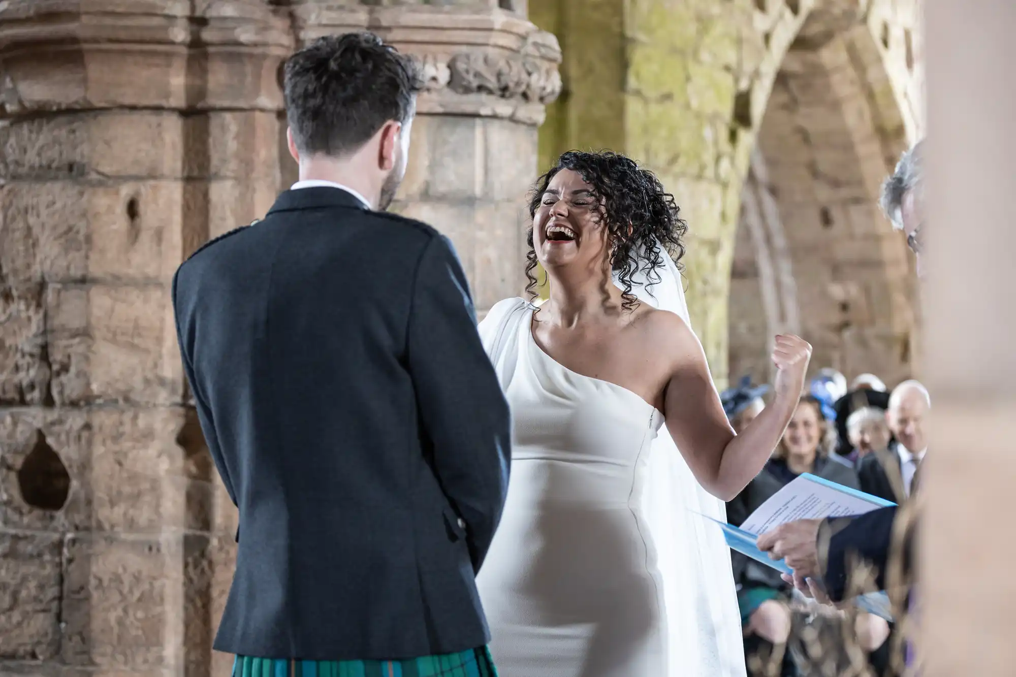 A bride in a white dress laughs while a groom in a kilt stands beside her during a wedding ceremony in an old stone building. Guests are seated in the background.