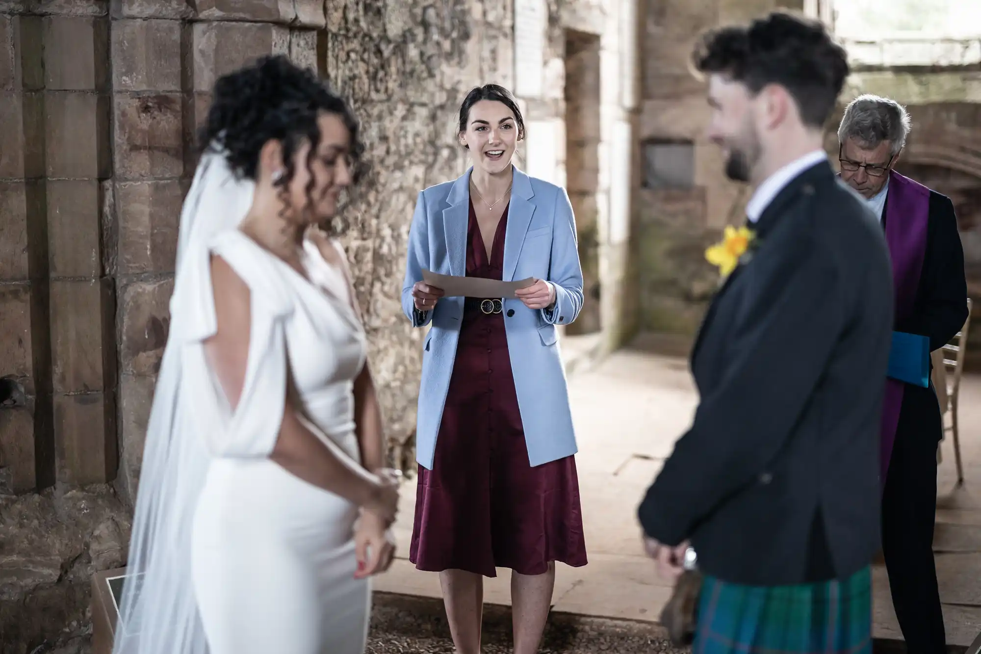 A wedding officiant stands between a bride in a white dress and a groom in a kilt inside a rustic stone building, reading from a document. Another person in a purple stole stands in the background.