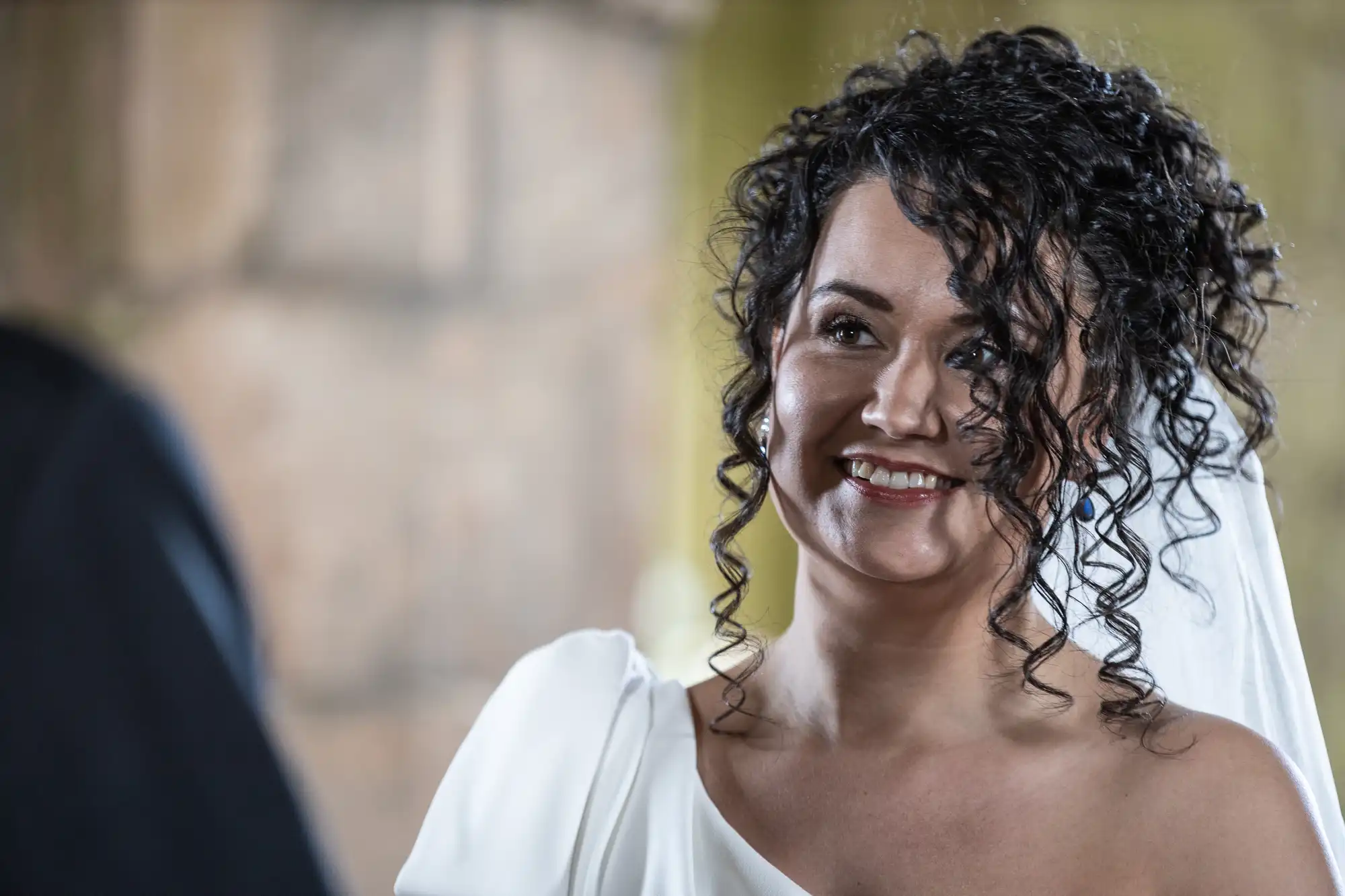 A smiling bride with curly hair and a white dress looks ahead. The background is blurred and rustic.