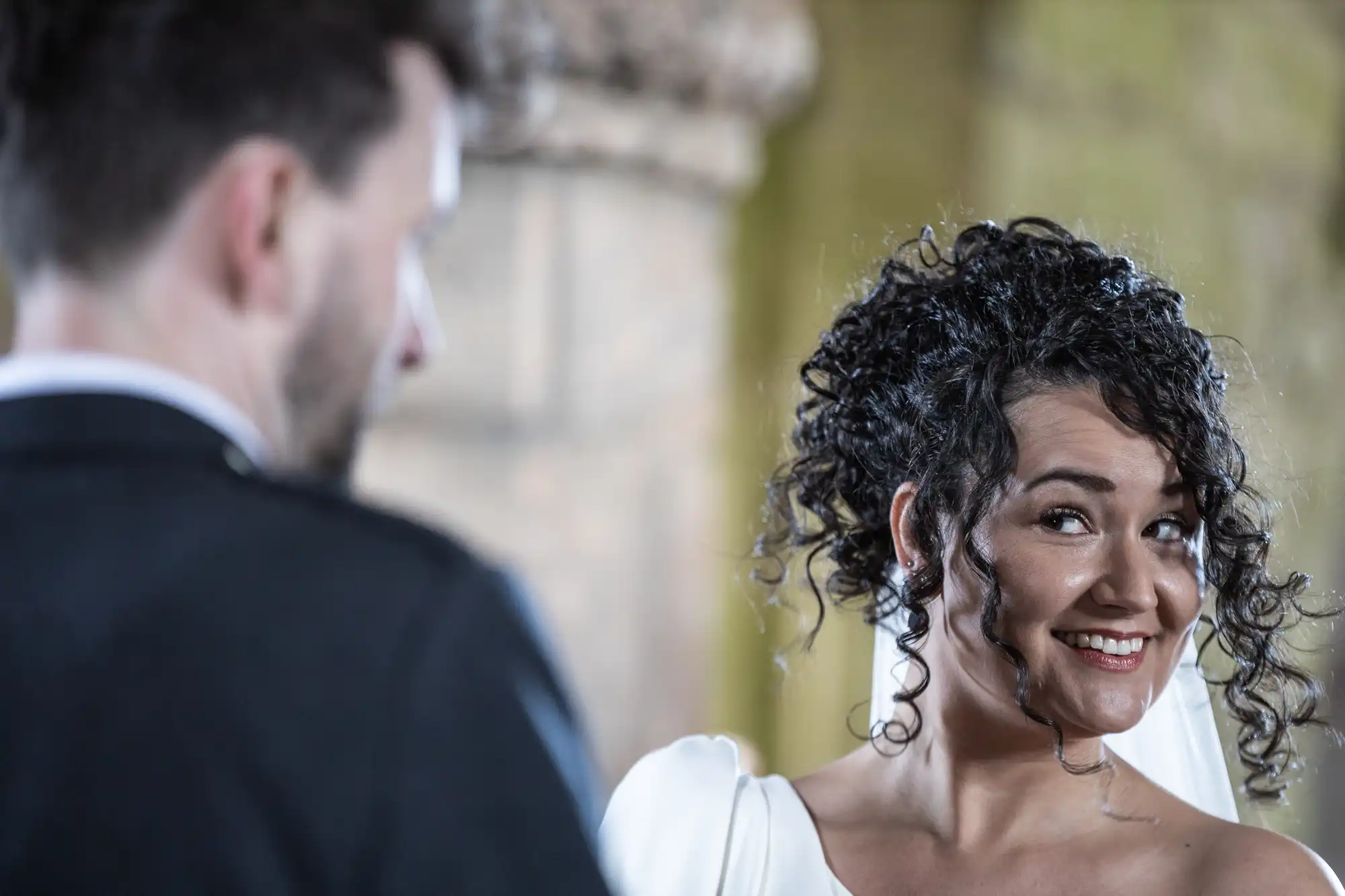 A woman with curly hair smiles while looking at a man with his back to the camera in an indoor setting with blurred background.