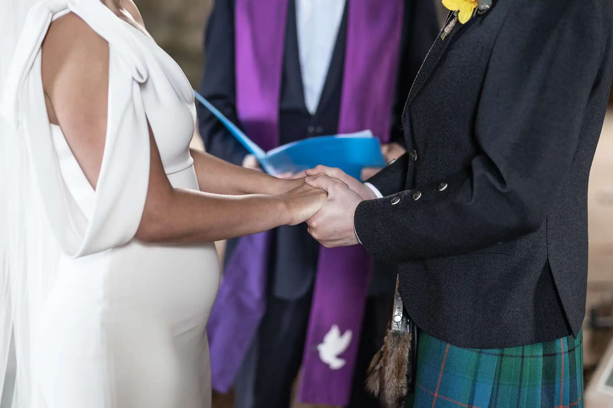 A bride and groom stand holding hands during a wedding ceremony officiated by a person in a purple stole. The bride wears a white dress and veil, and the groom is in a dark suit with a tartan kilt.