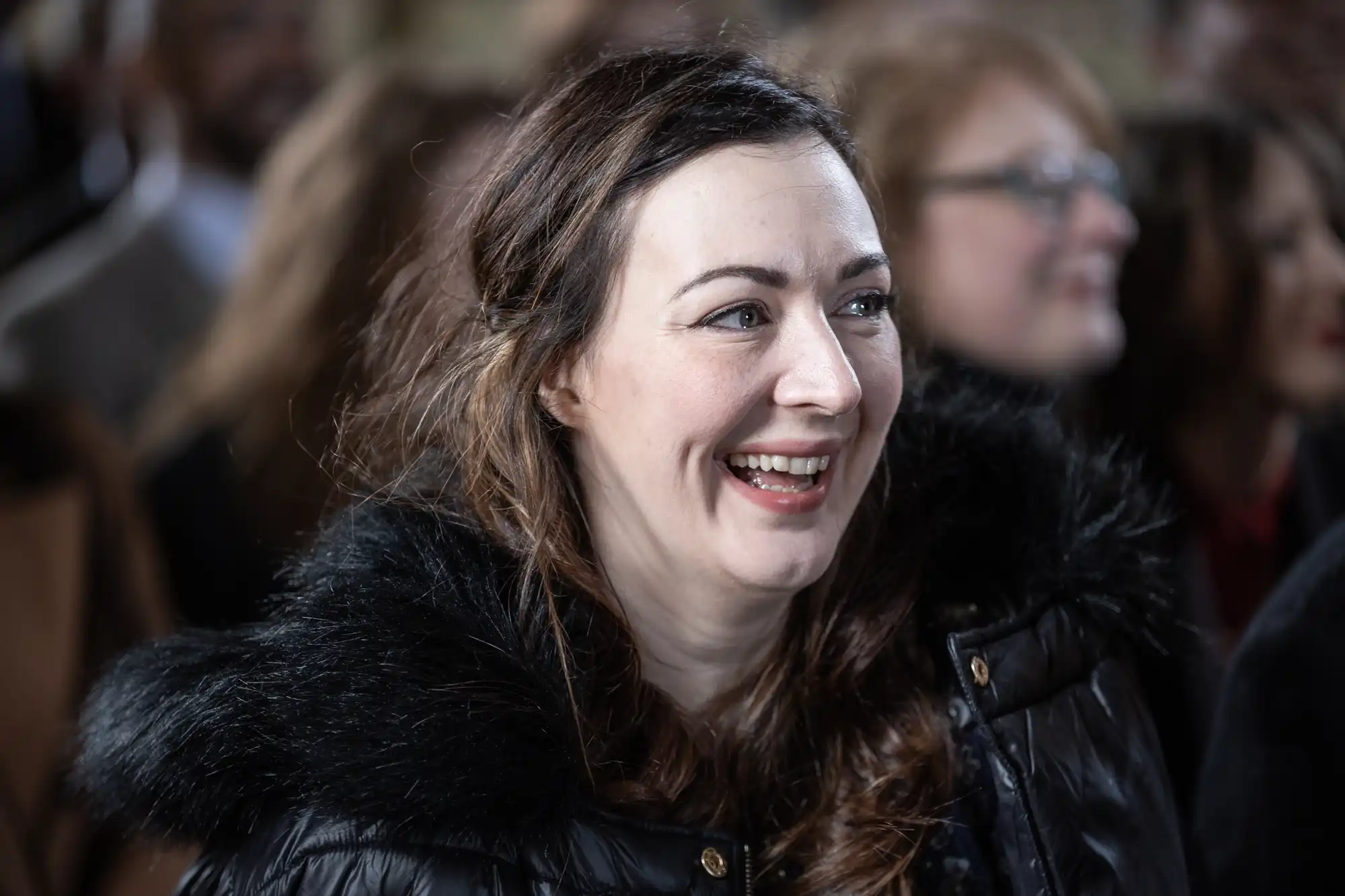 A woman with brown hair, wearing a black coat with a fur collar, smiles in a crowd.