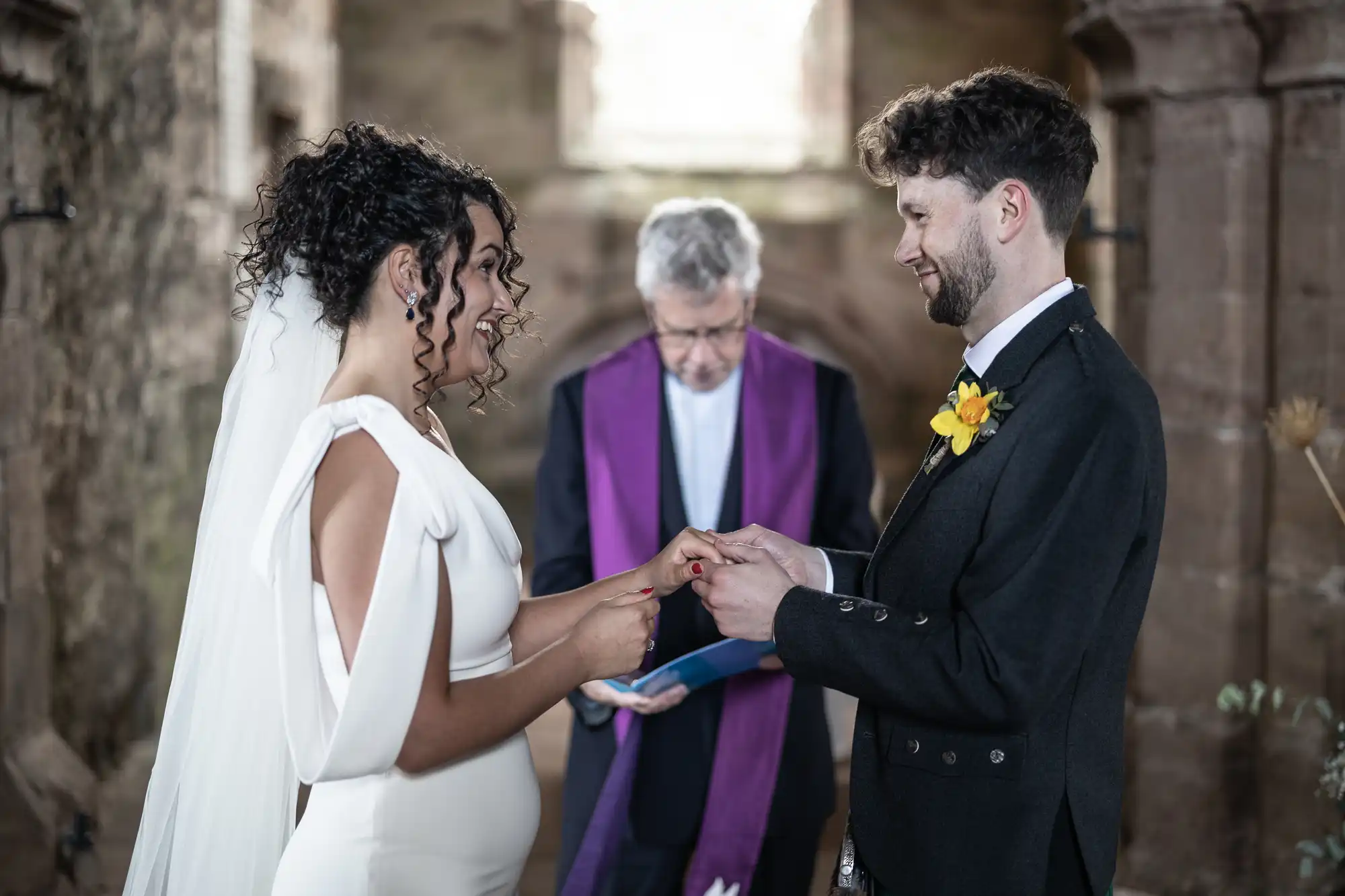 A bride and groom exchange rings in a stone building during their wedding ceremony, with an officiant in a purple stole standing behind them.