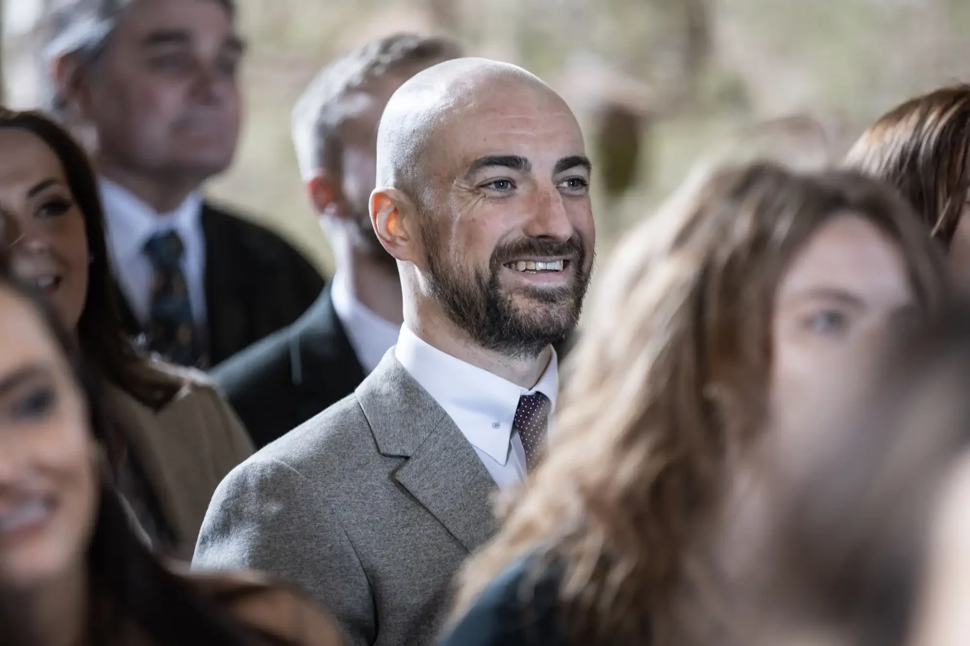 A man with a beard and a bald head, wearing a gray suit, smiles while standing in a crowd during an unplugged wedding ceremony