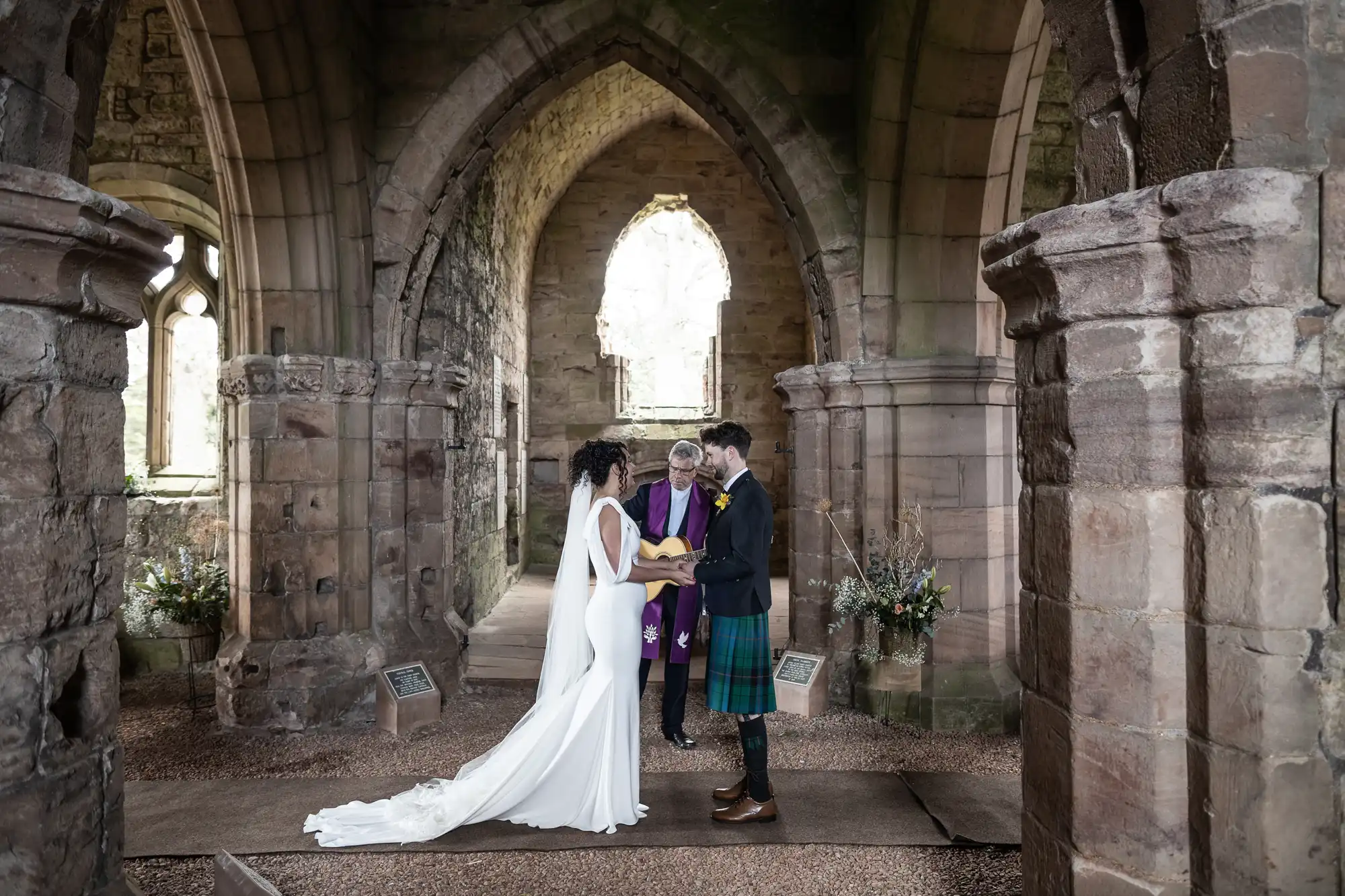 A couple stands holding hands during a wedding ceremony inside an old stone church, with a person officiating in the background.